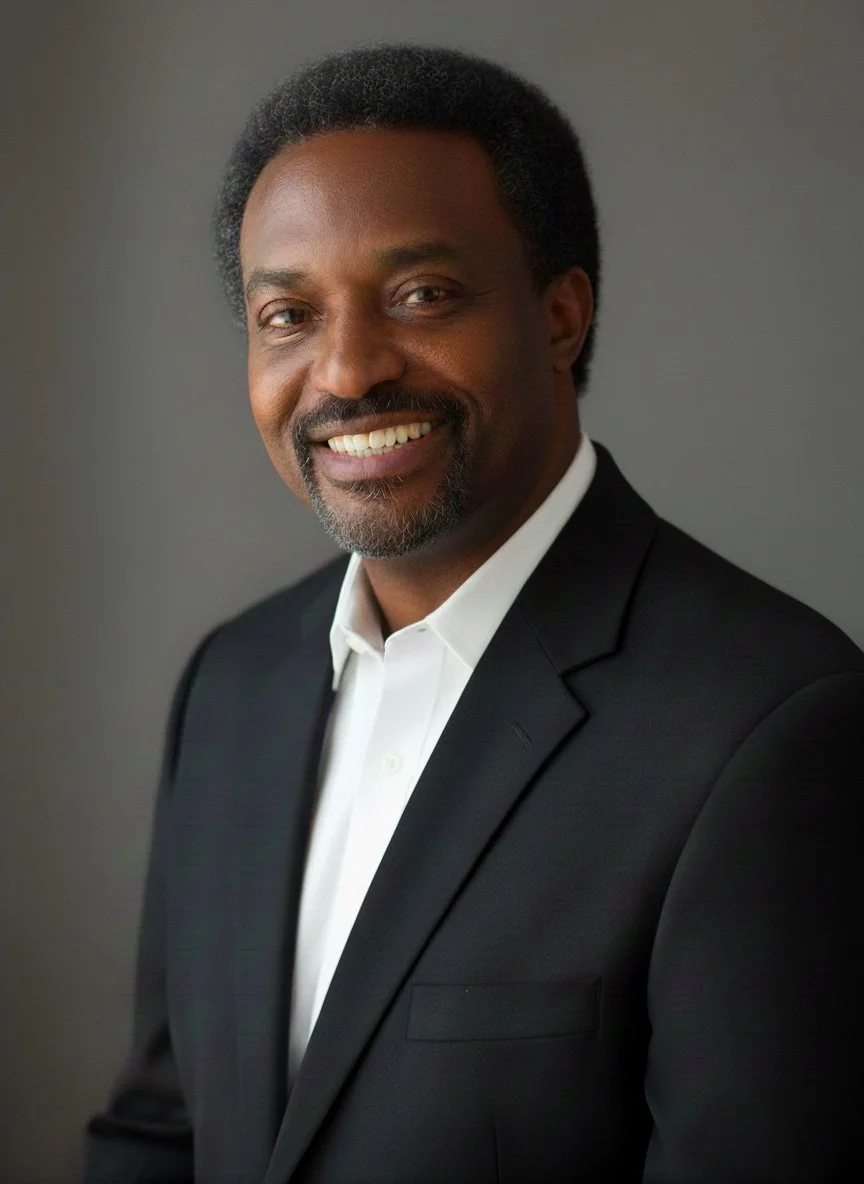 A portrait of a smiling African American man wearing a black suit and white shirt against a gray background.