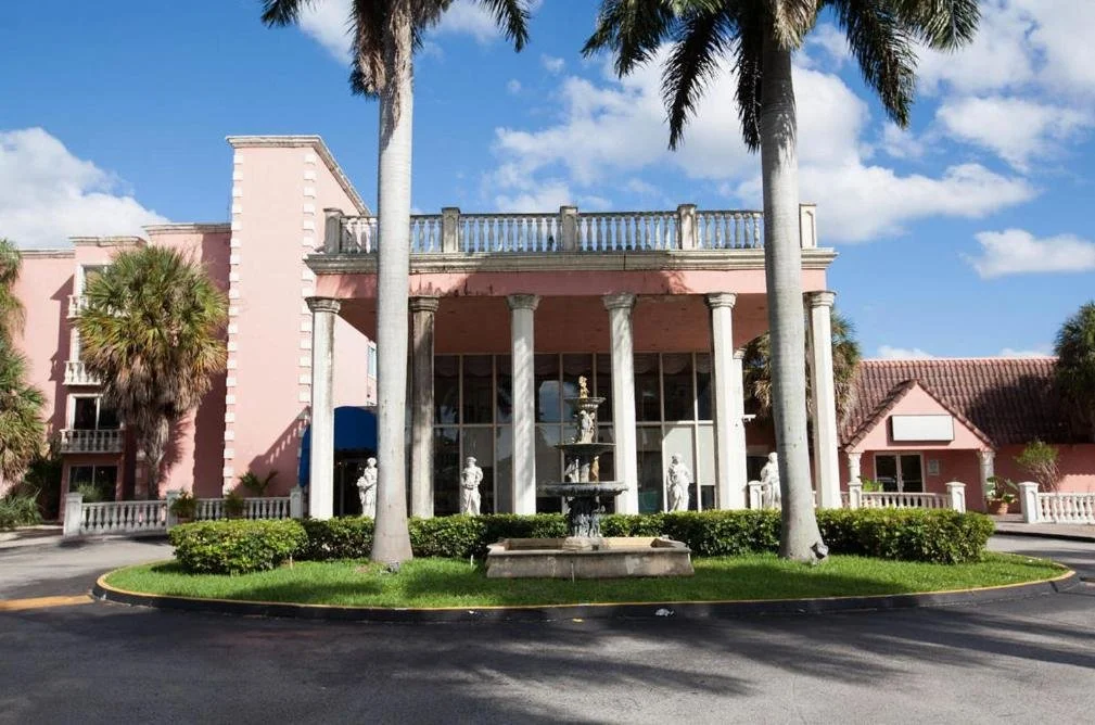 A pink building with classical columns, statues in front, palm trees, and a fountain in the center under a partly cloudy sky.