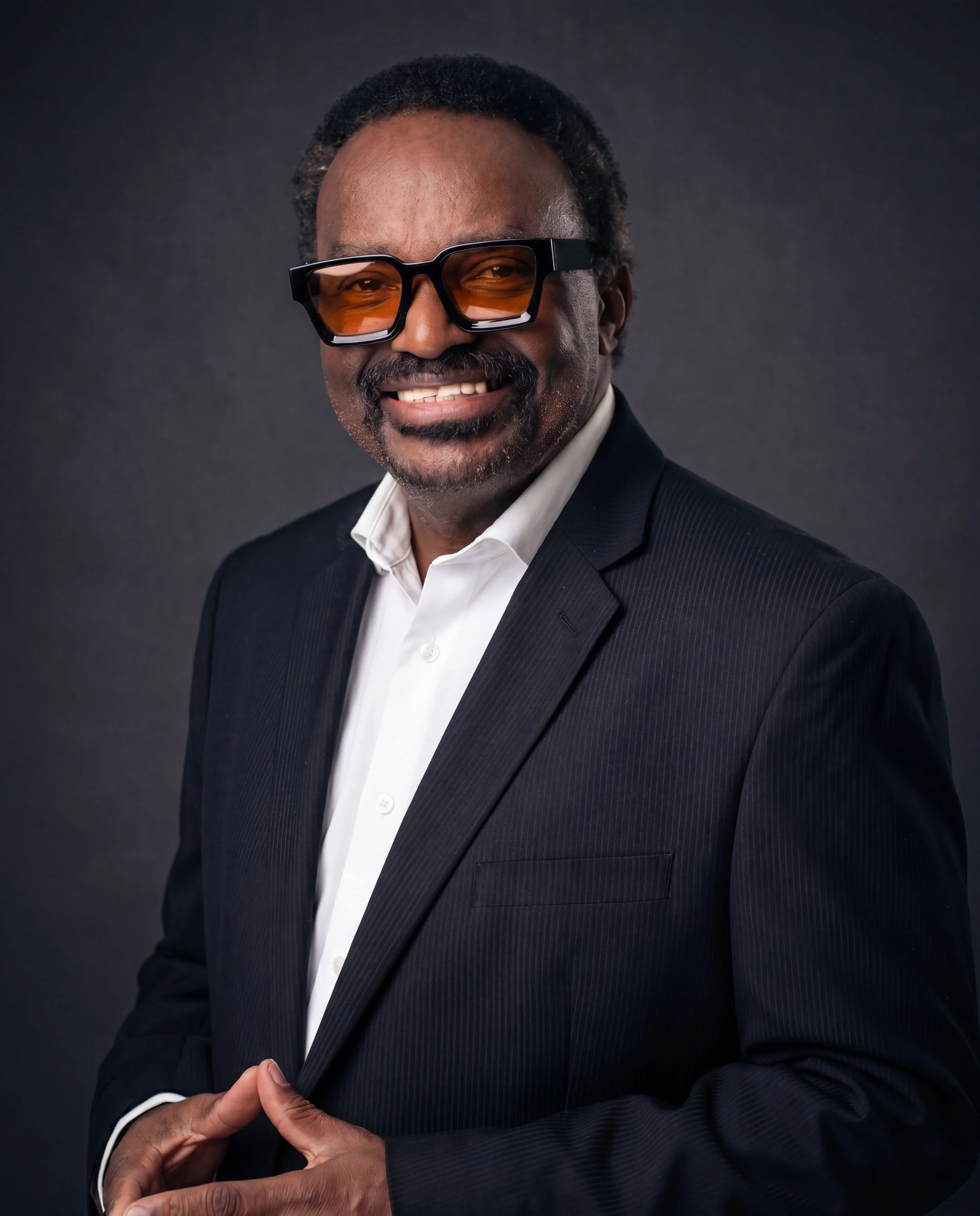 A portrait of a smiling African American man wearing a black suit and white shirt against a gray background.