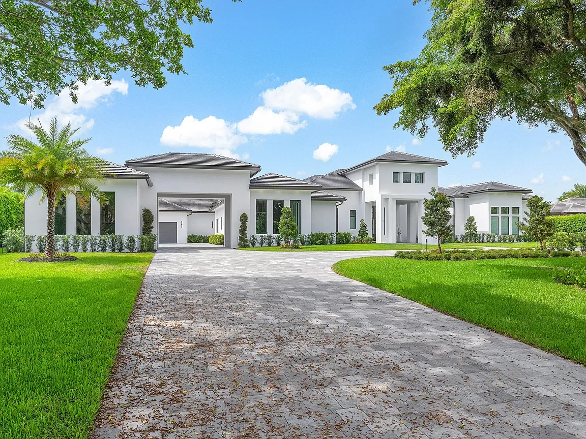 Large white modern house with a paved driveway, surrounded by green grass and trees, under a blue sky with white clouds.