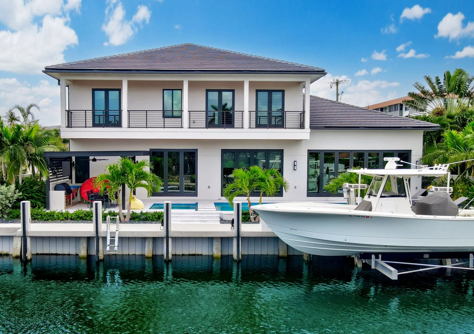 Modern two-story house by water with boat, pool, and palm trees