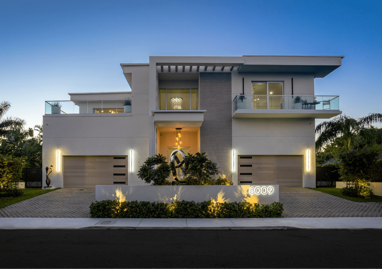 Modern white multi-story house with balconies, illuminated entrance, landscaped front yard with plants and sculpture, house number 8009, during early evening.