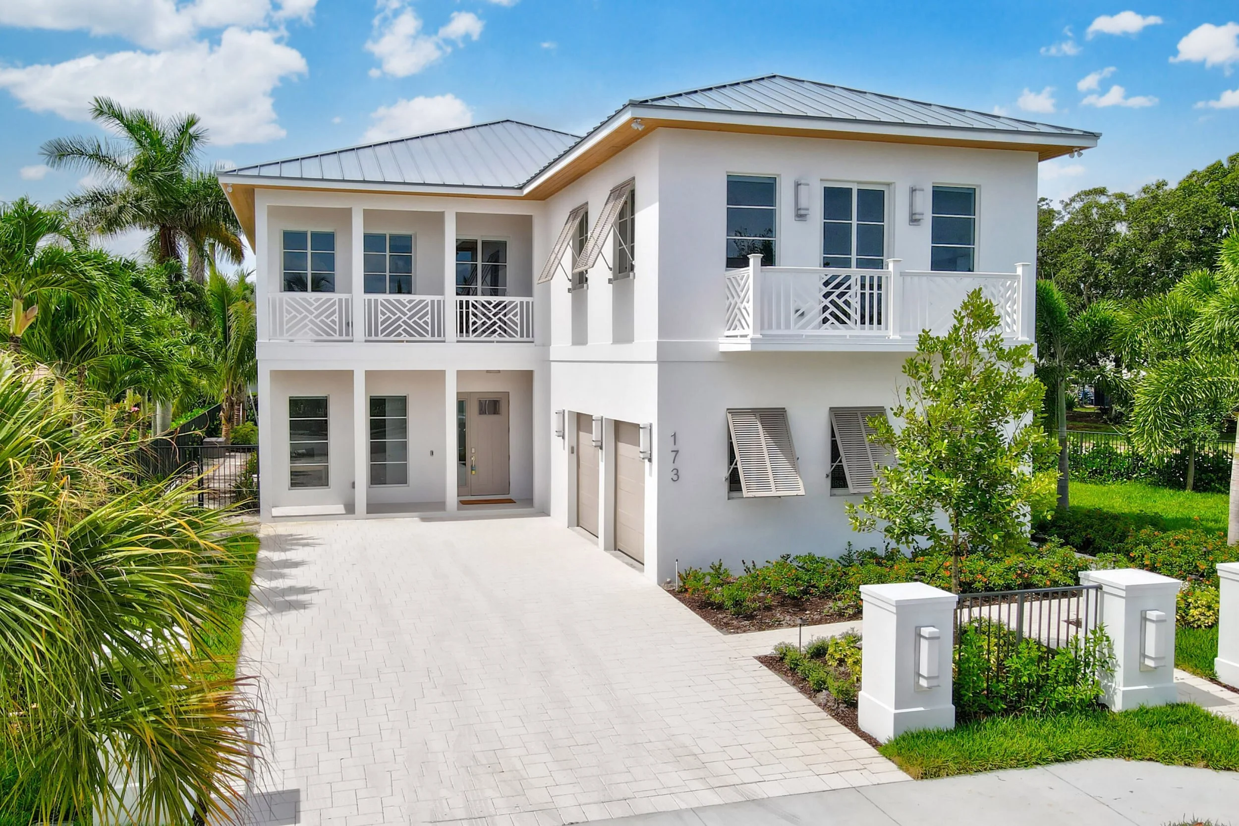 Modern two-story white house with gray roof, surrounded by tropical plants and a white paved driveway.