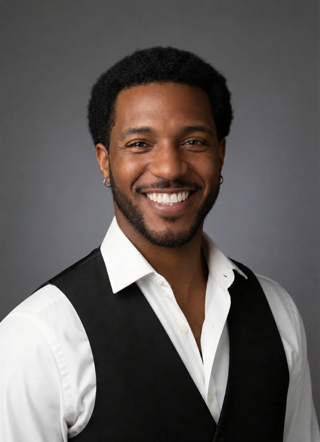 A smiling man with dark curly hair, wearing a white shirt and black vest, standing against a gray background.