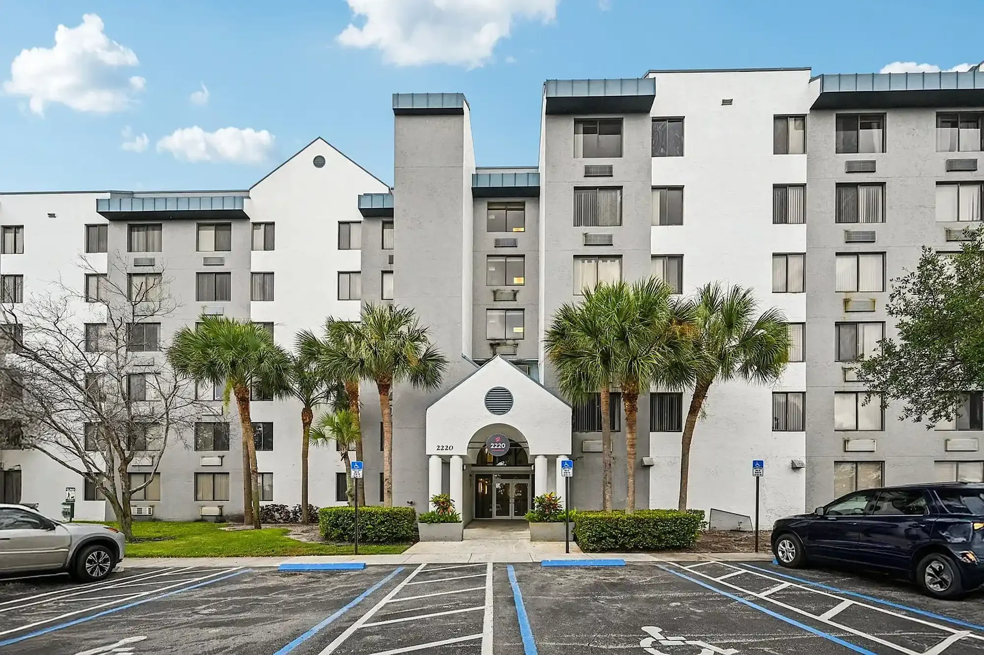 A white multi-story apartment building with palm trees in front, office blinds visible through some windows, and a parking lot with handicapped and regular parking spaces