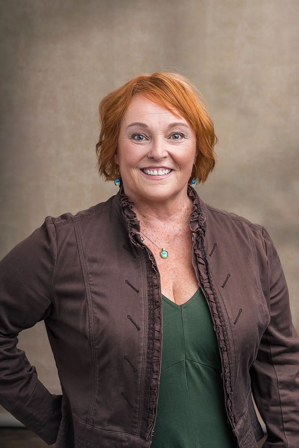 Smiling woman with short red hair wearing a brown jacket and green top, against a neutral background.