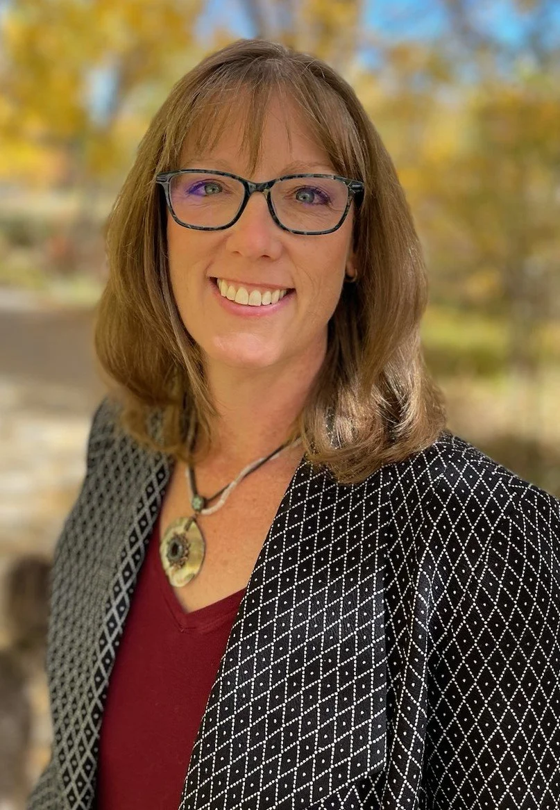 Woman with glasses smiling outdoors in patterned jacket and necklace, autumn background.