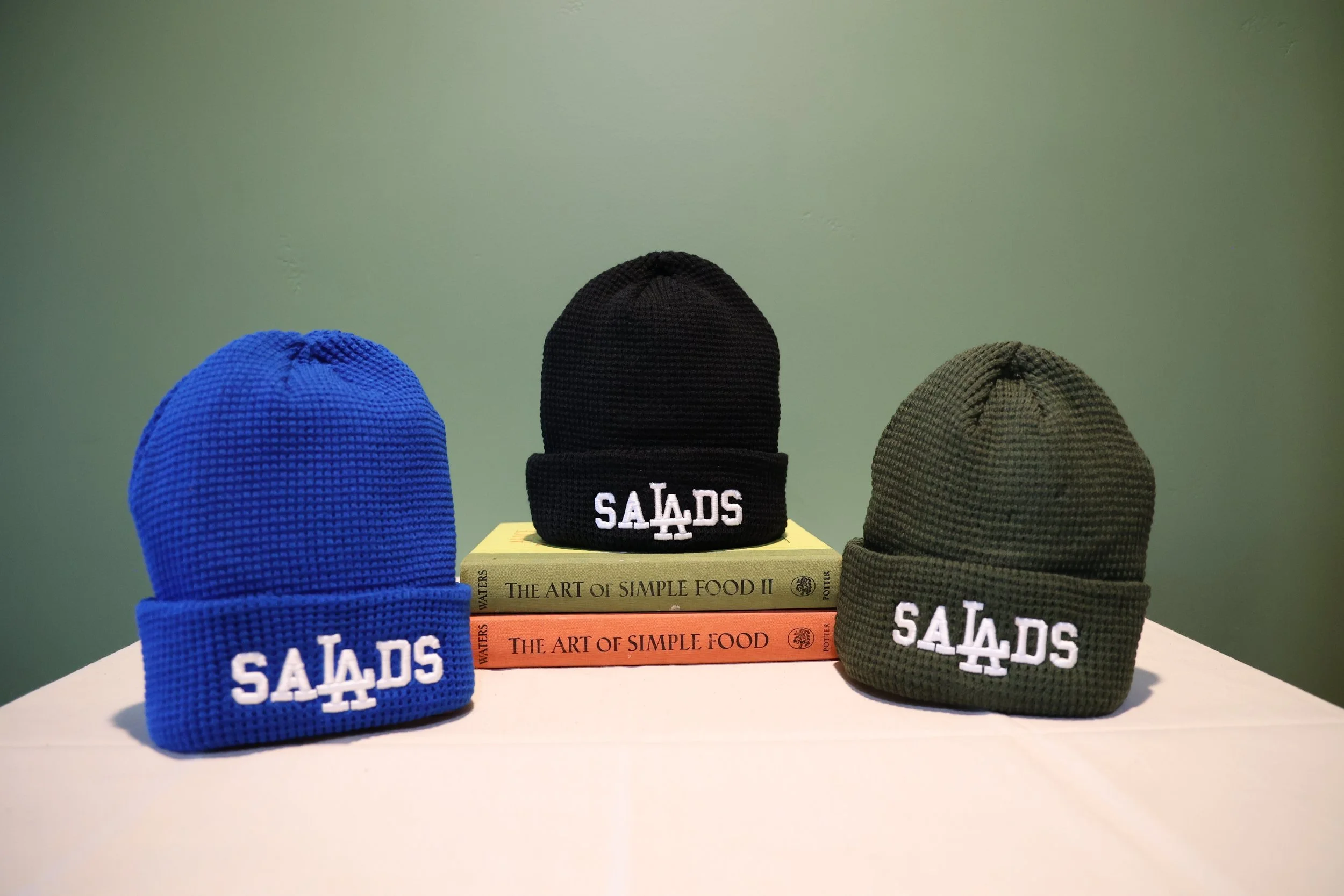 3 comfy beanies in blue, black and green that read SALADS on the forehead