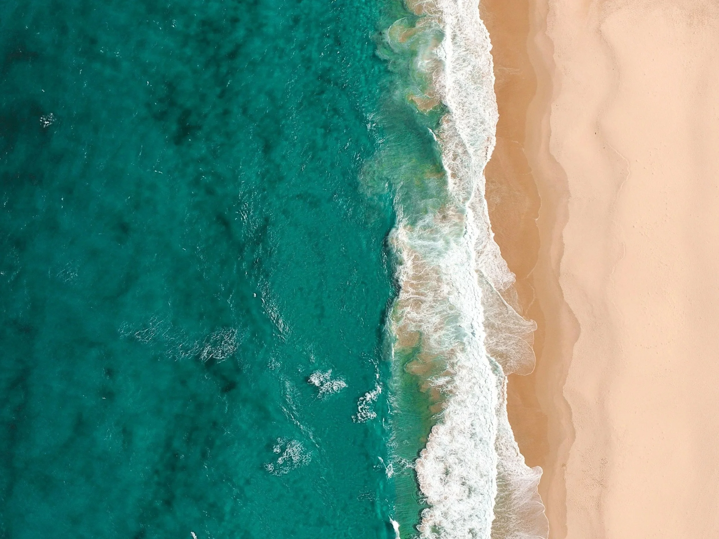 Aerial view of bright turquoise ocean meeting pale golden sand, with white waves rolling onto the shore. The kind of summer break that only works when your freelance business is set up to run without you.