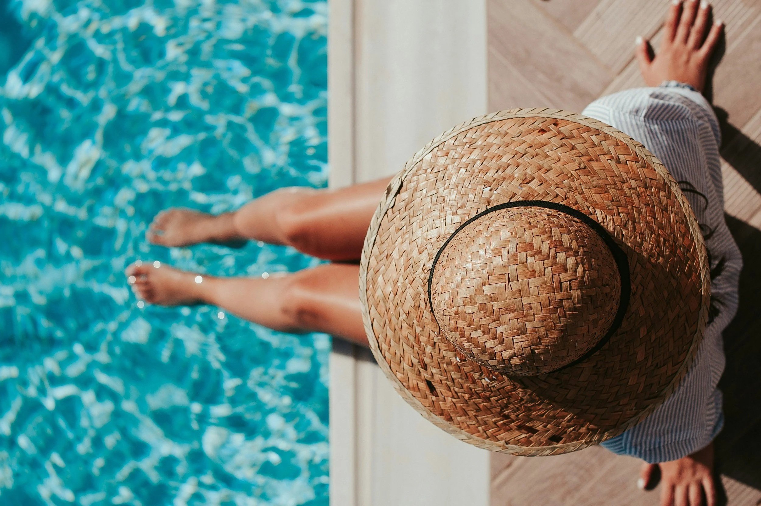 Over-the-shoulder view of someone in a wide-brimmed straw sun hat, sitting at the edge of a swimming pool with bare legs and feet dipped into bright turquoise water. The unplugged summer moment this post is about.