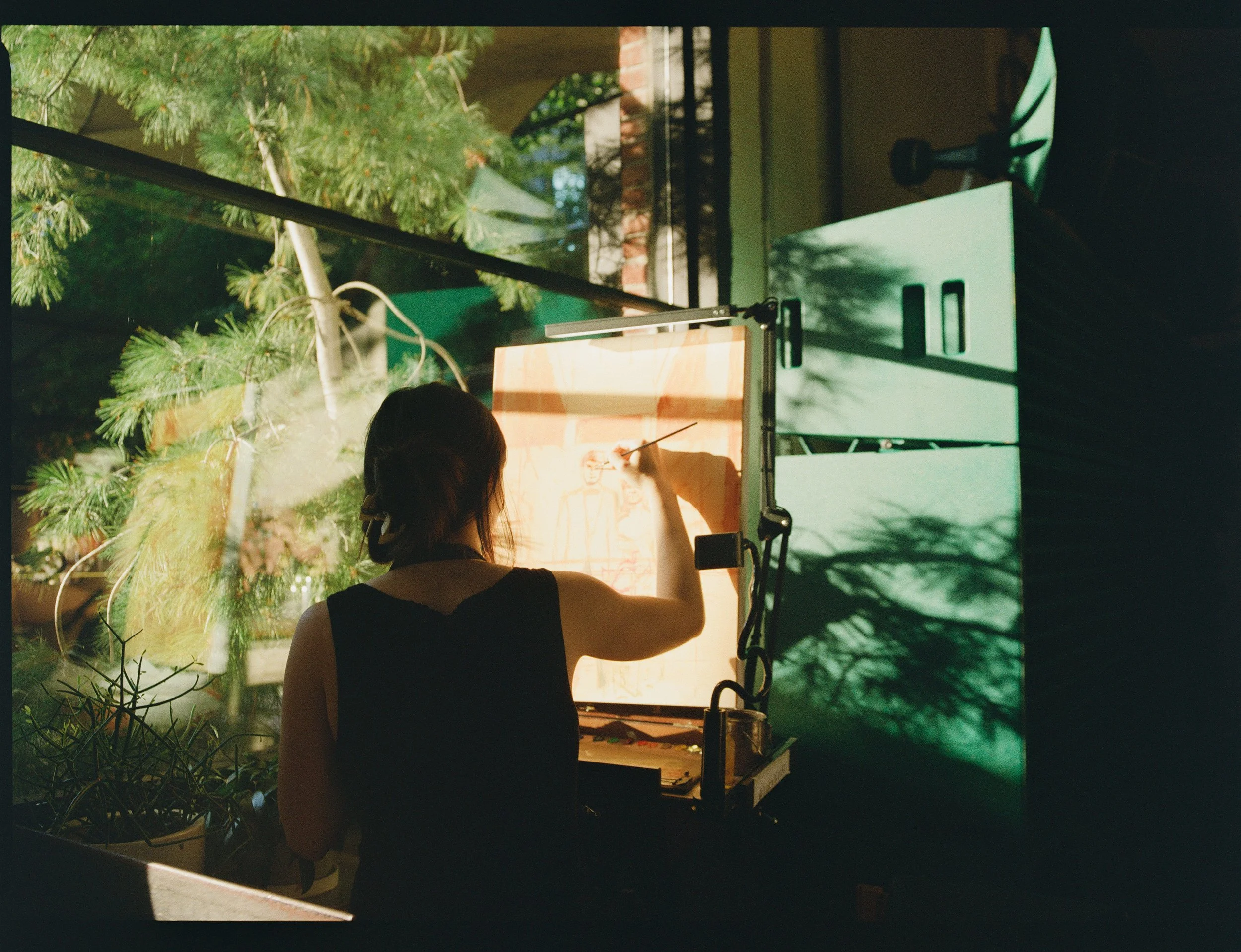 A woman painting a portrait on a bright lightbox with sunlight streaming through a large window and green plants outside.