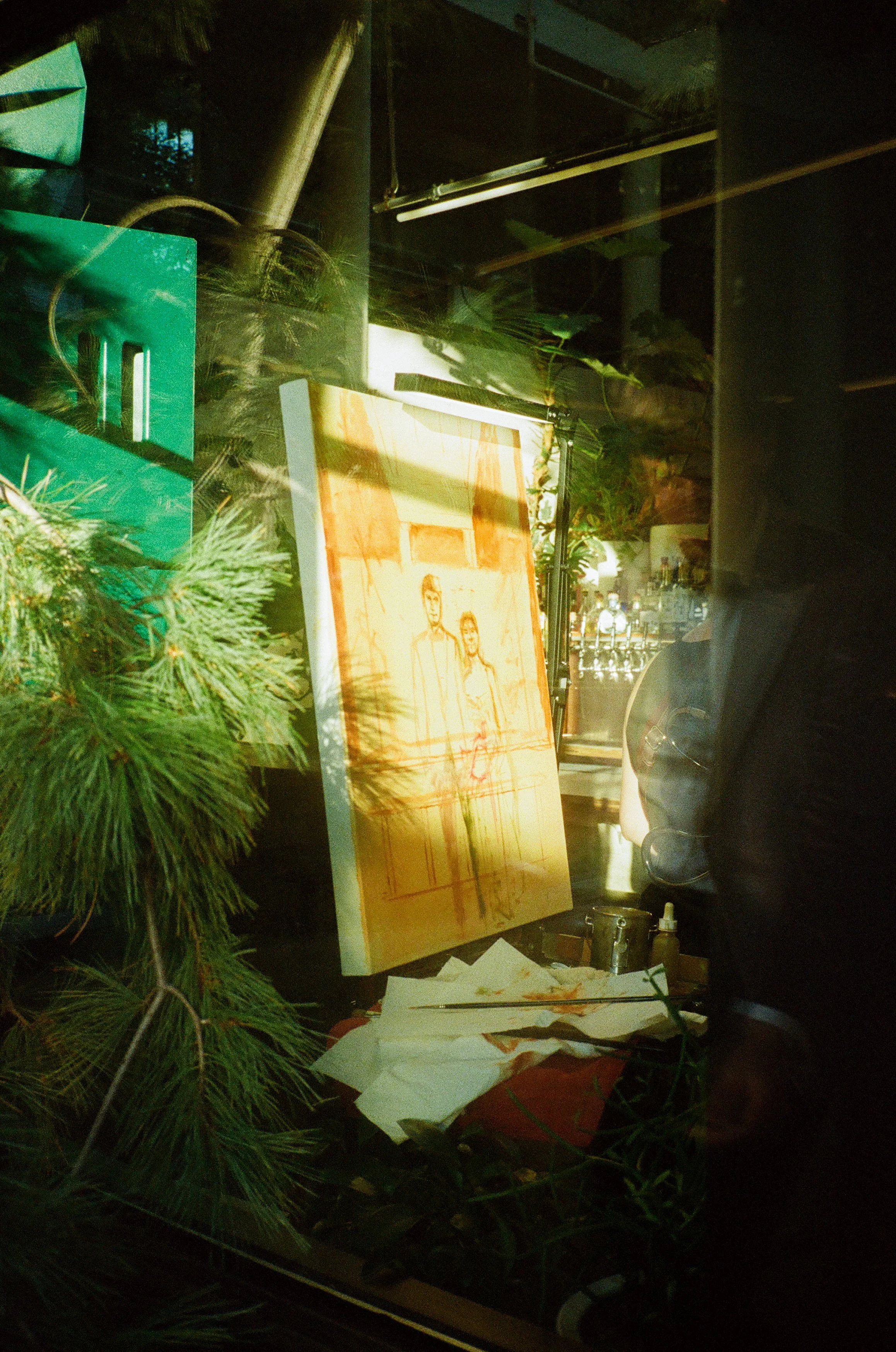A window display with a painting of two people, some greenery, art supplies, and a bar with bottles reflected in the glass.