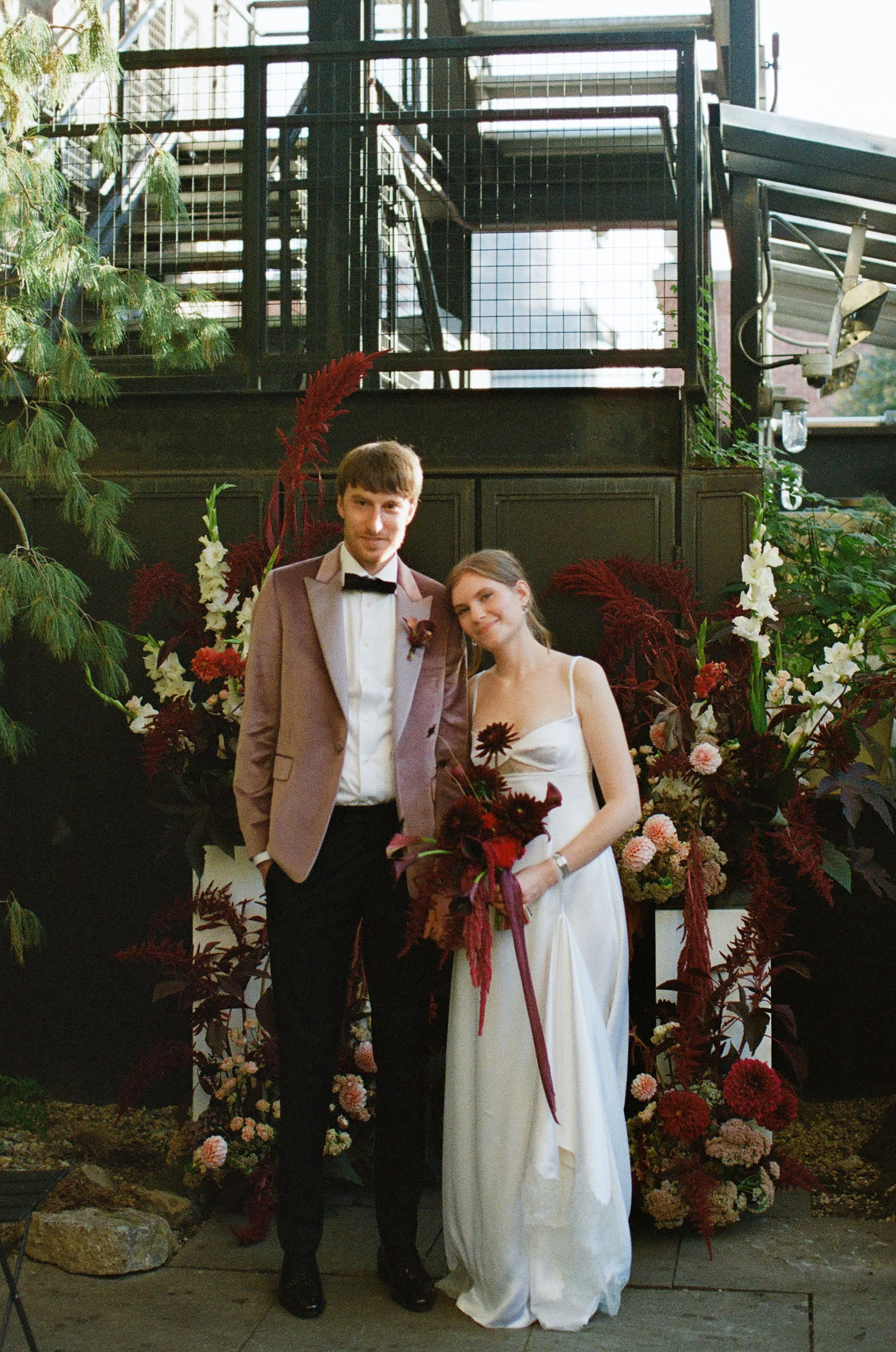 A couple dressed in formal wedding attire standing in front of a large floral display featuring dark red and white flowers and greenery.