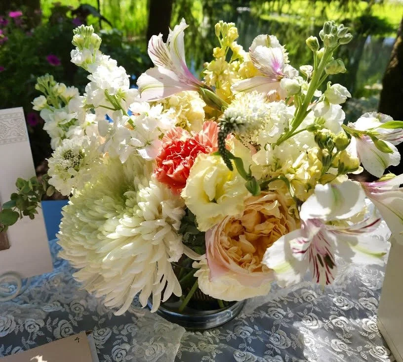 A floral arrangement in a black vase with various white, pink, and yellow flowers, including lilies, carnations, and dahlias, placed on a table covered with a patterned cloth outside in a garden setting.