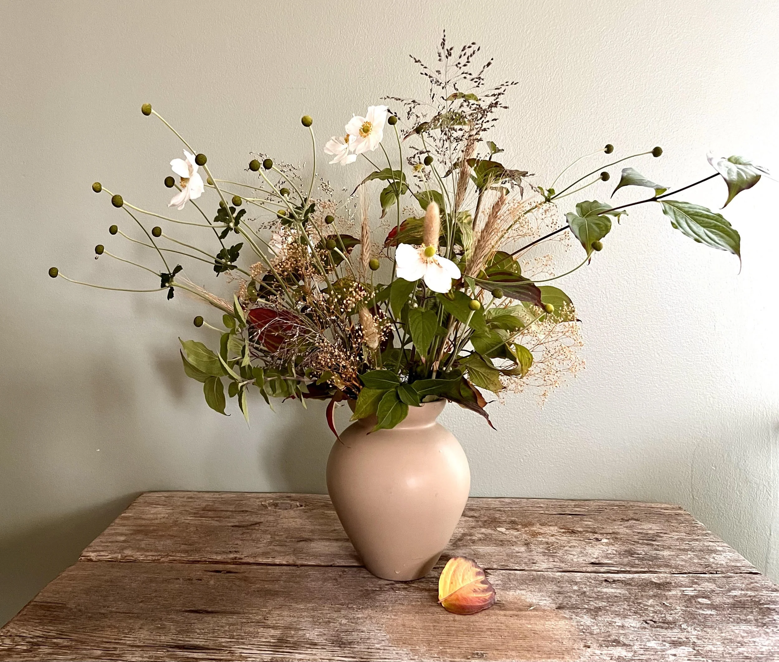 A beige ceramic vase filled with a mixed bouquet of flowers and greenery sits on a rustic wooden table, with a fallen petal in front of it.