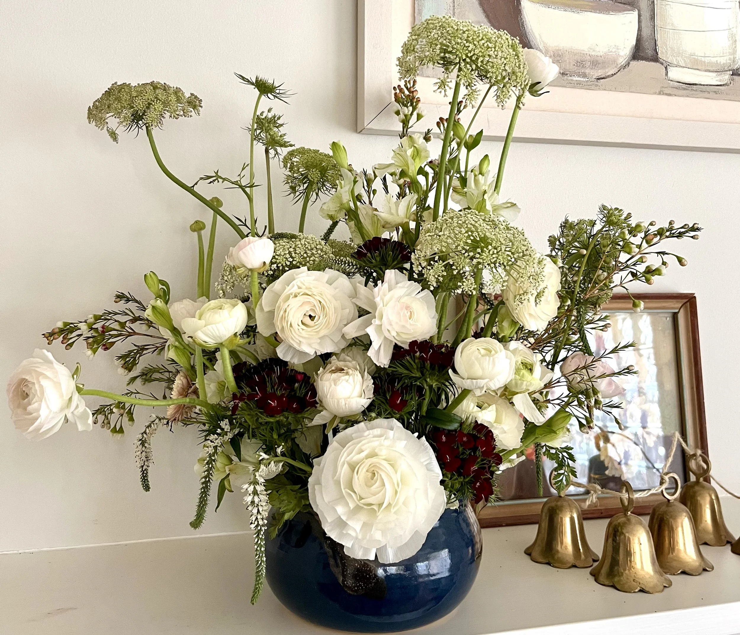 A bouquet of white and green flowers in a navy ceramic vase on a white surface behind some gold-colored decorative objects, with a framed abstract artwork hanging on a white wall in the background.