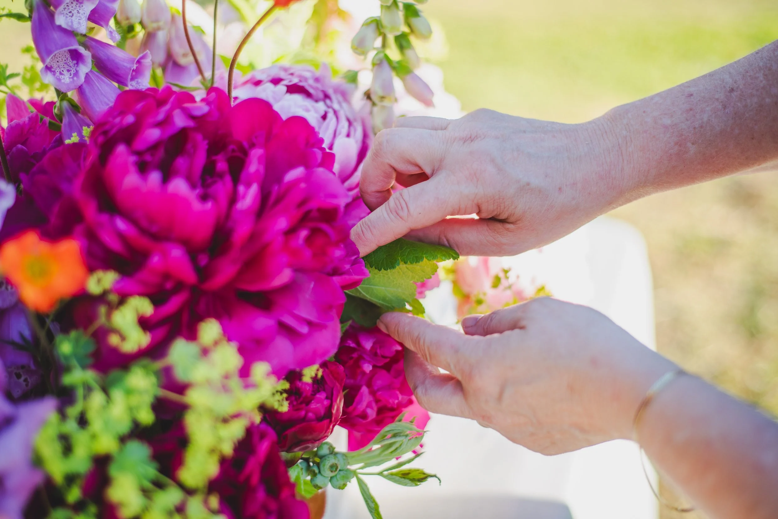 A person's hands arranging a bouquet of pink, purple, and yellow flowers outdoors on a sunny day.