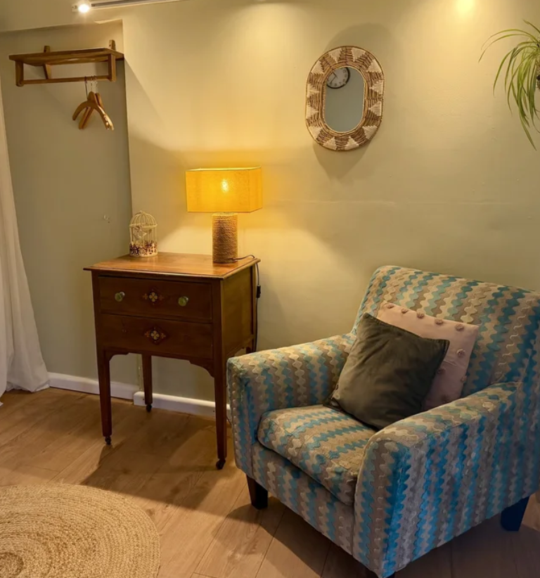 Cozy living room corner with an upholstered armchair, a wooden side table with a yellow lamp, a mirror on the wall, and decorative pillows.