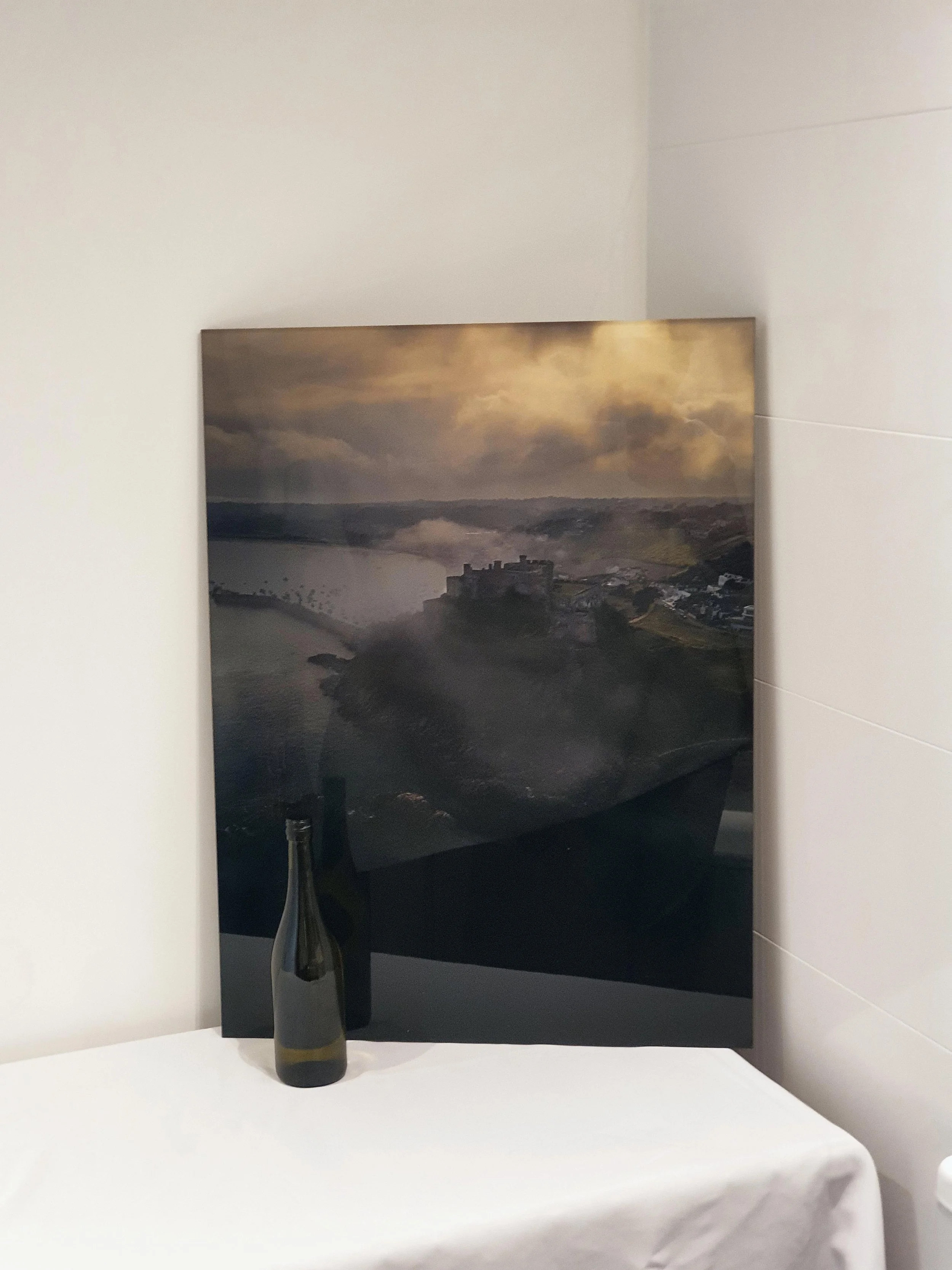 A black bottle with a gold and white label, placed on a white table in front of a framed landscape photograph showing a river, a castle, and cloudy sky.