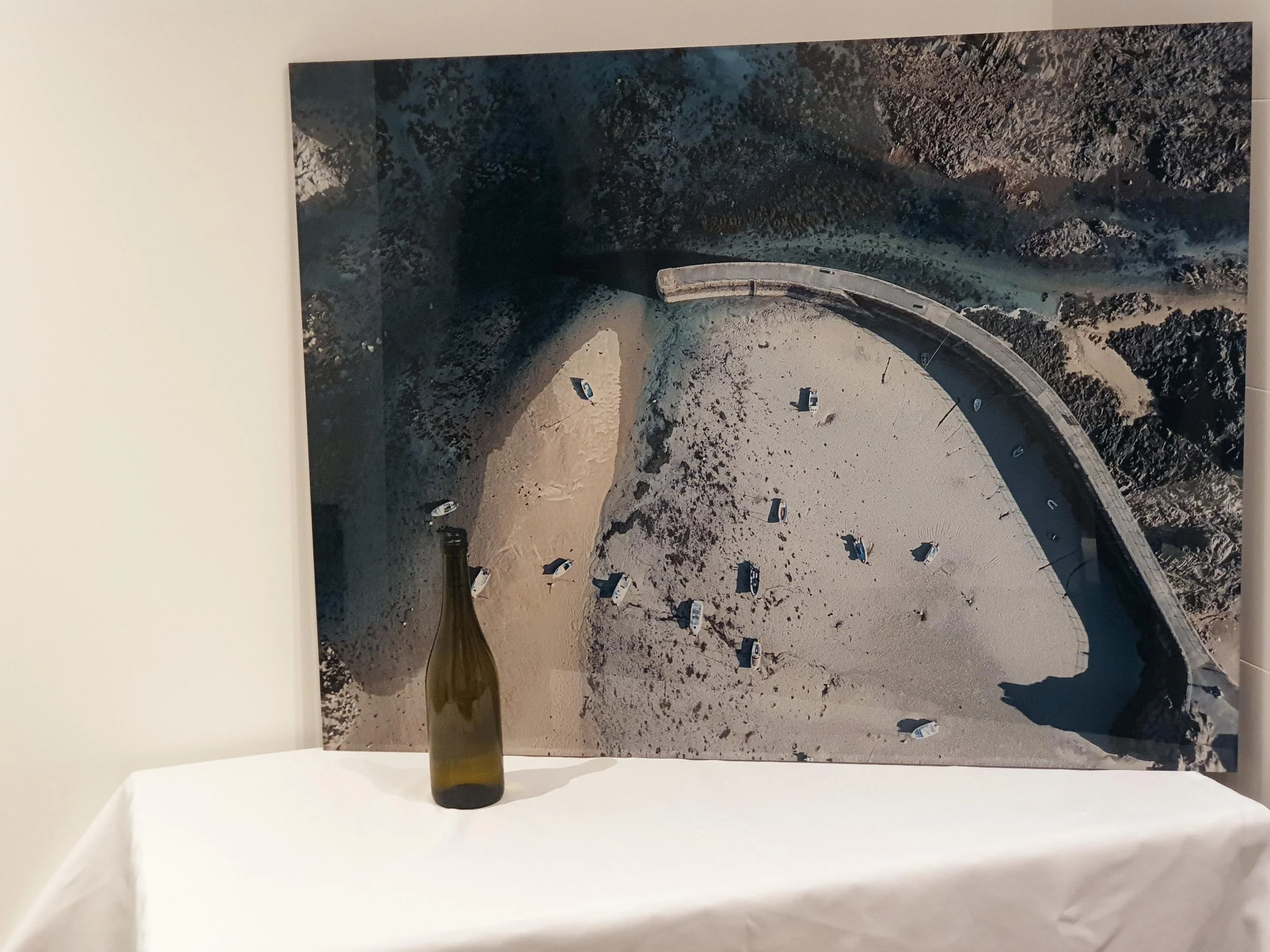 A photograph of a white gallery wall featuring a large aerial landscape photograph with a winding road, parked cars, and rugged terrain. The photo is mounted on the wall, and a dark green-greenish bottle is placed on a white table below it.