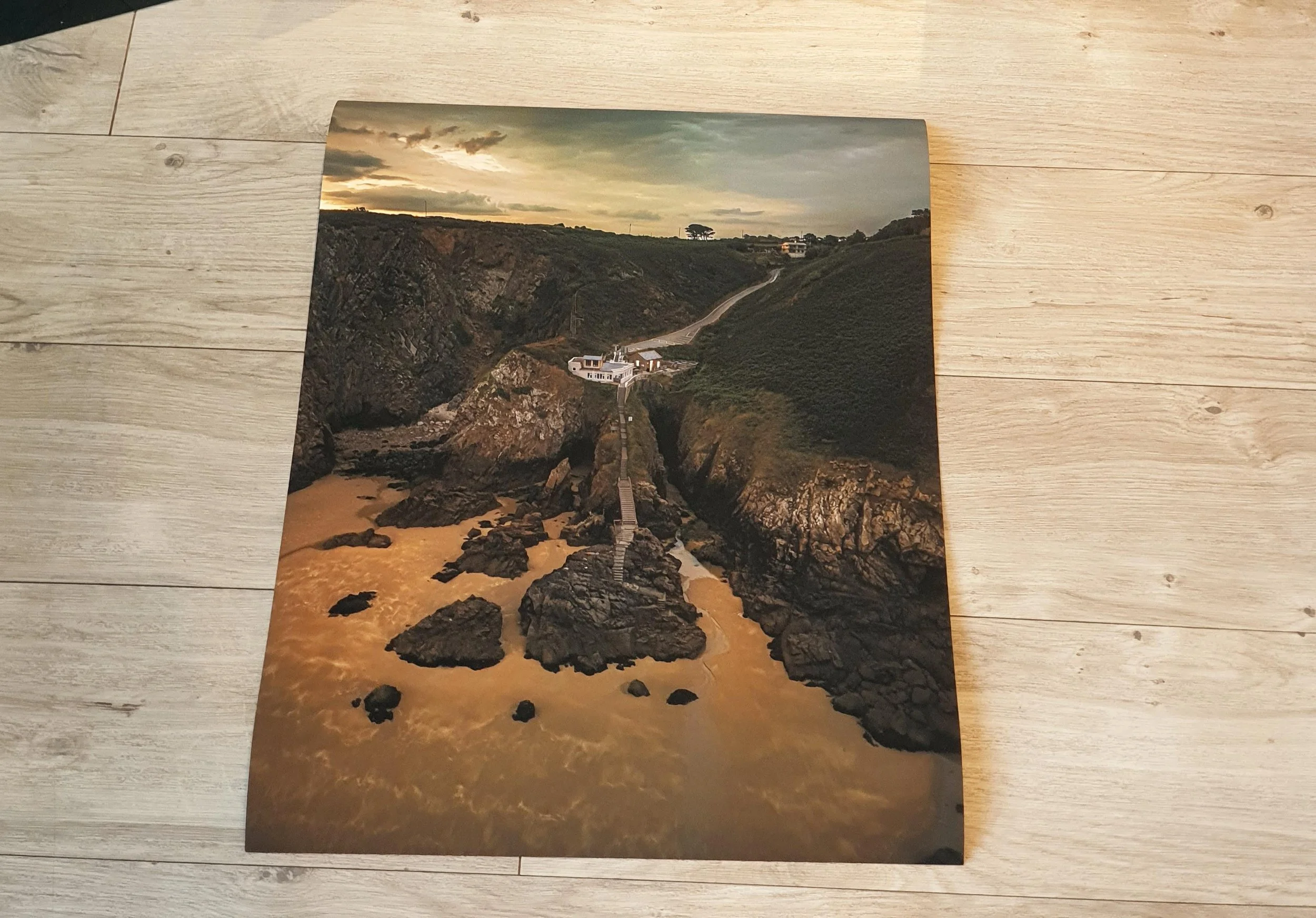 A scenic photograph of a coastal landscape showing a rocky shoreline, a beach, and a pathway leading to houses on a hillside under a cloudy sky at sunset.