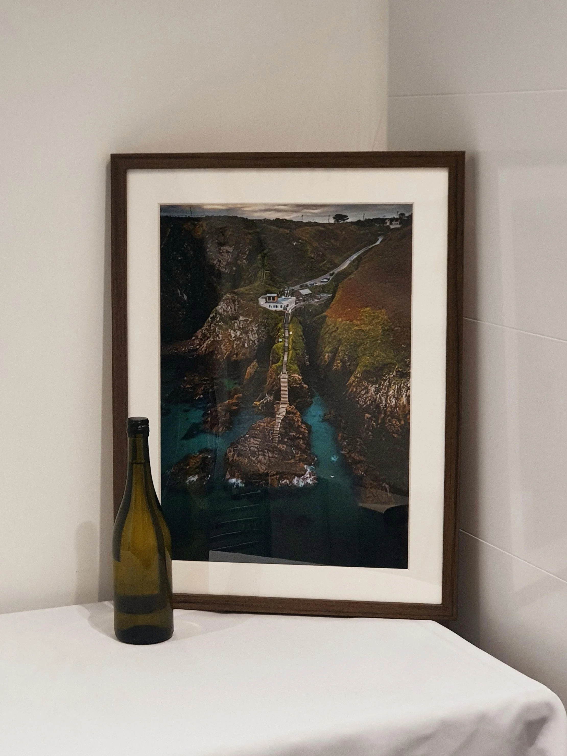 A framed landscape photograph of a coastal scene with a walkway bridge leading to a house on a hill, next to water, with a wine bottle in front of the frame on a white surface.