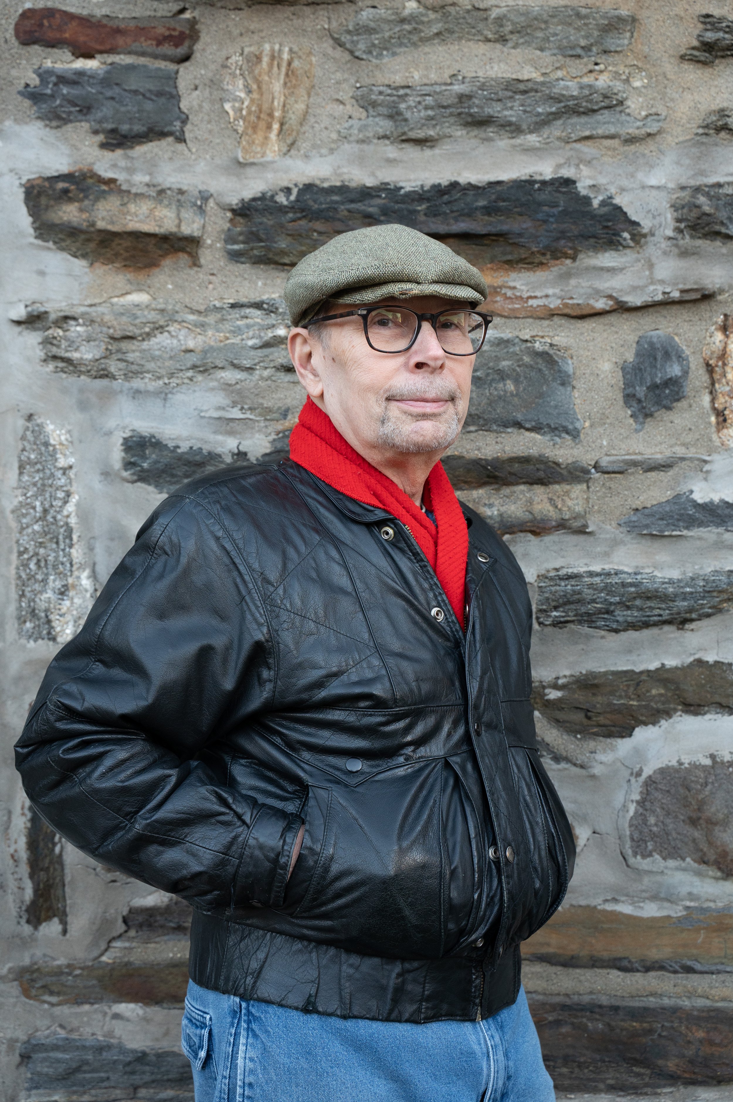 Eugene DeGeorge stands in front of stone wall in a leather jacket and red scarf