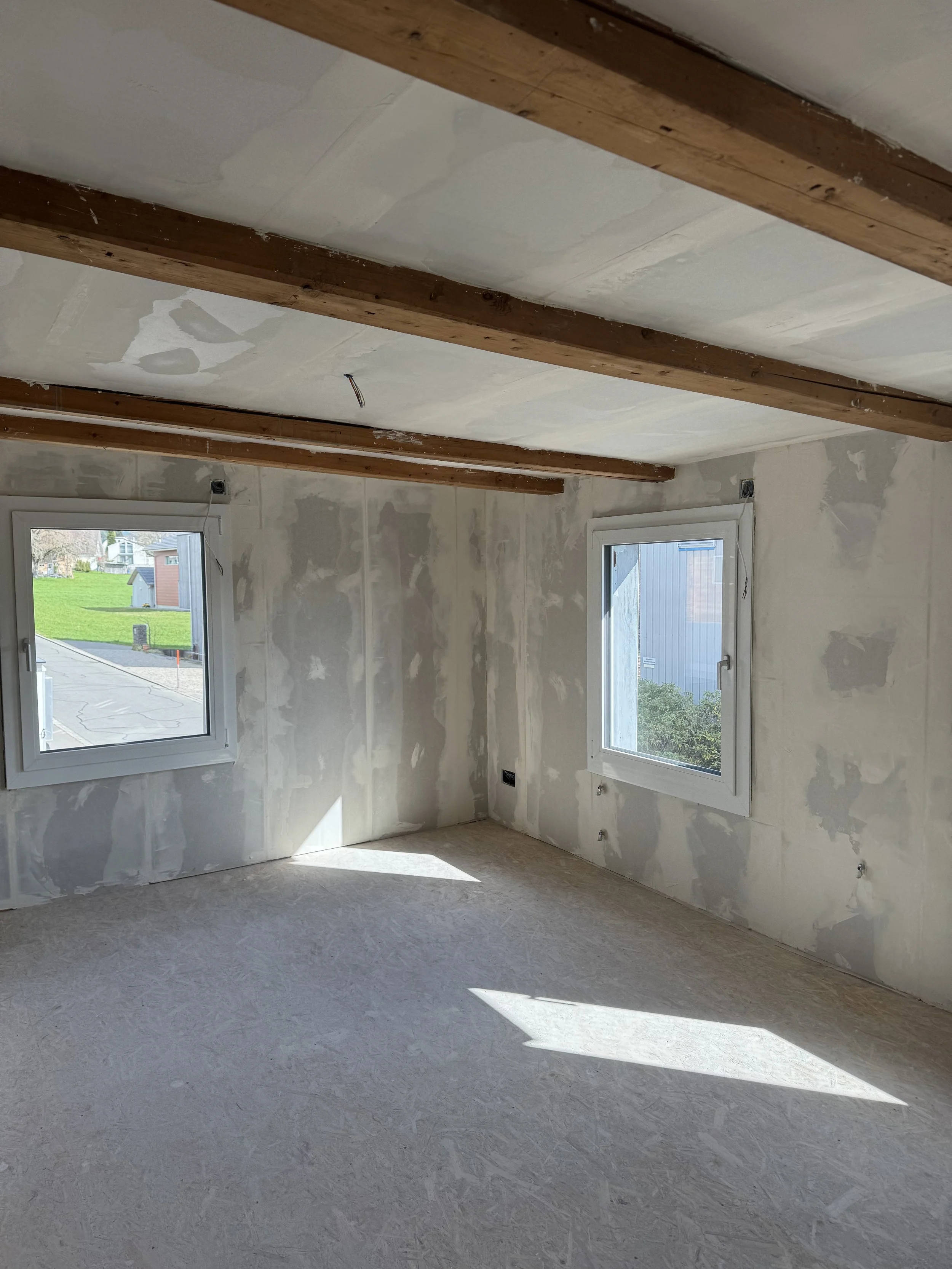 Empty room under construction with unfinished walls, two windows, and exposed wooden beams on the ceiling.