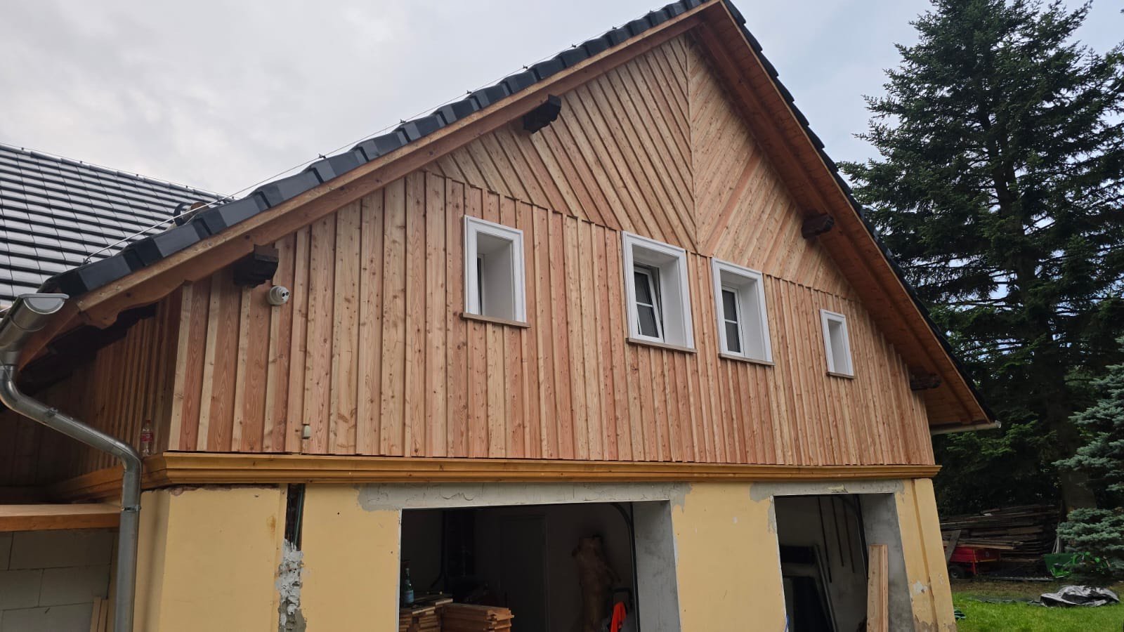 Side view of a two-story house under construction, with new wooden siding and small rectangular windows on the upper floor. The house has a sloped roof with dark gray shingles, and there is construction work along the bottom part of the house.