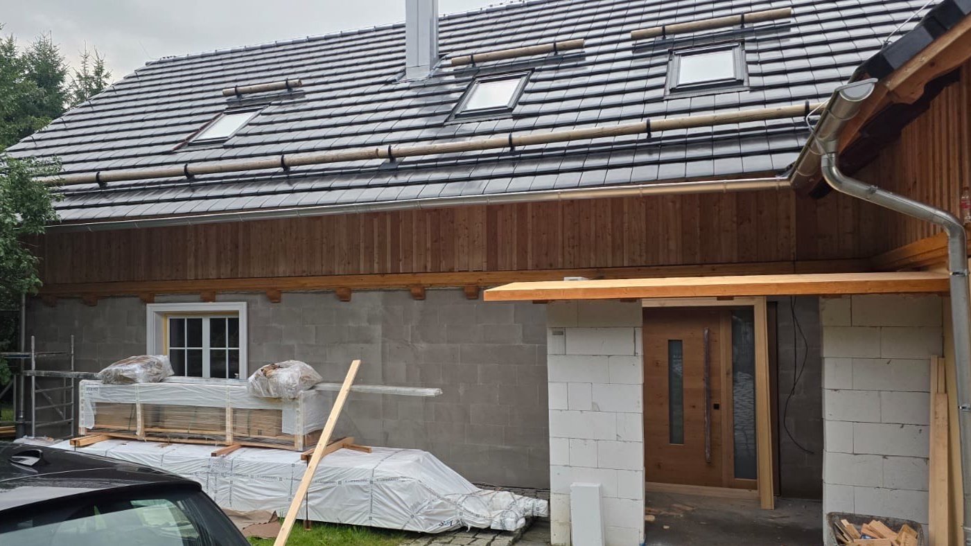 House under construction with gray tiled roof, wooden and brick walls, skylights, and construction materials in front.