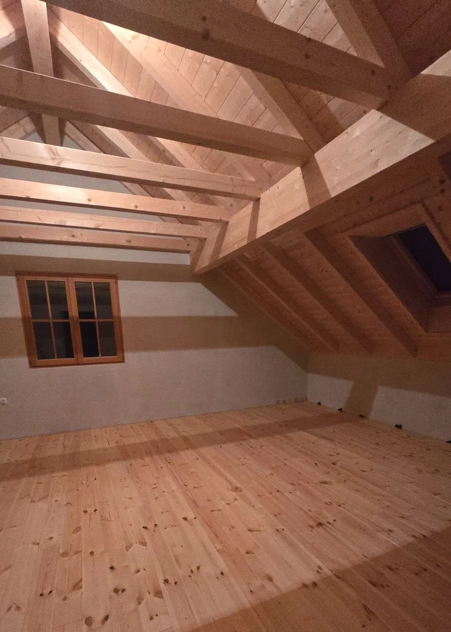 Empty attic room with wooden walls, floor, and ceiling beams, and a small window on the wall.