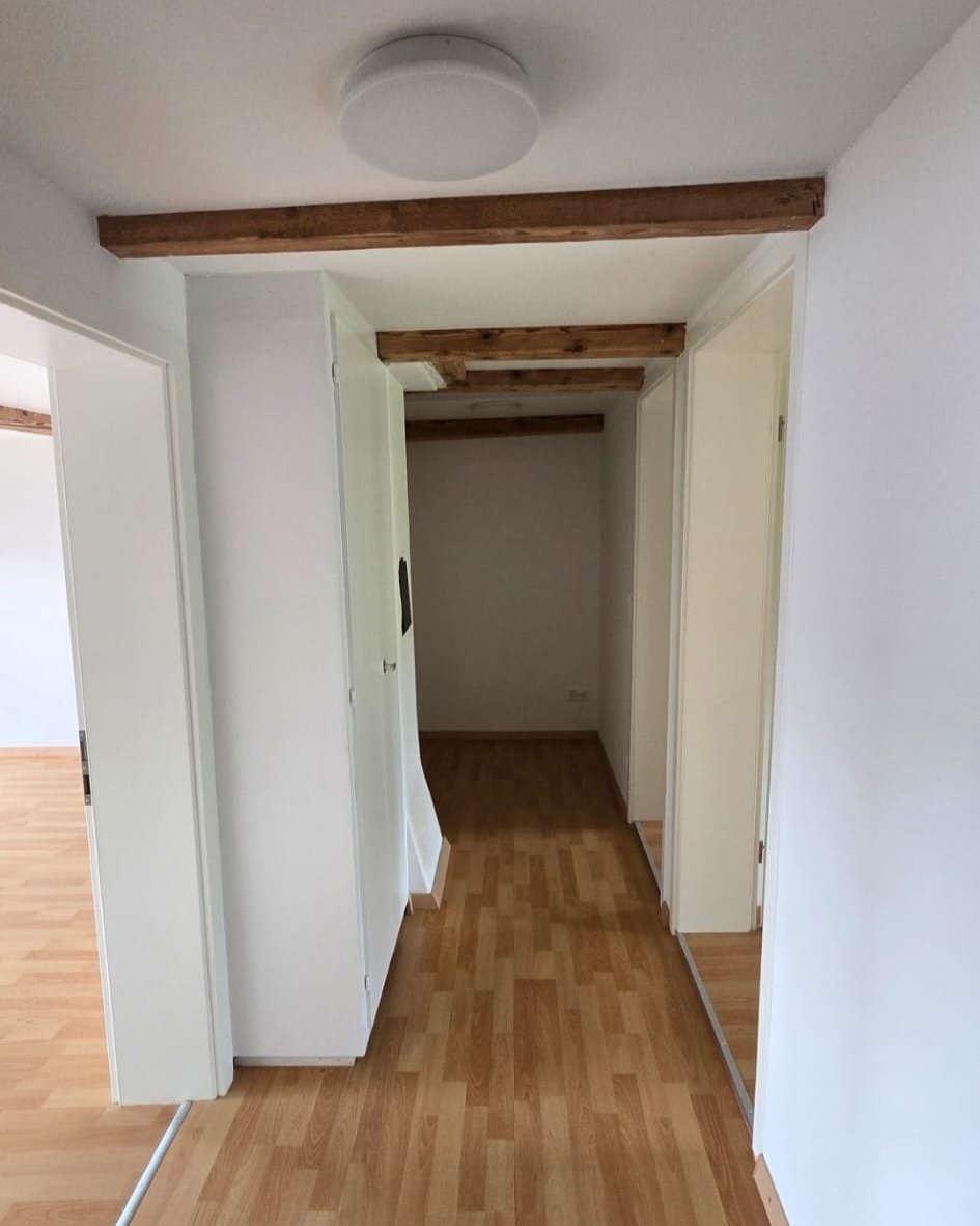 View of a hallway with unfinished ceiling beams and white walls, leading into a small recessed area with wooden flooring.