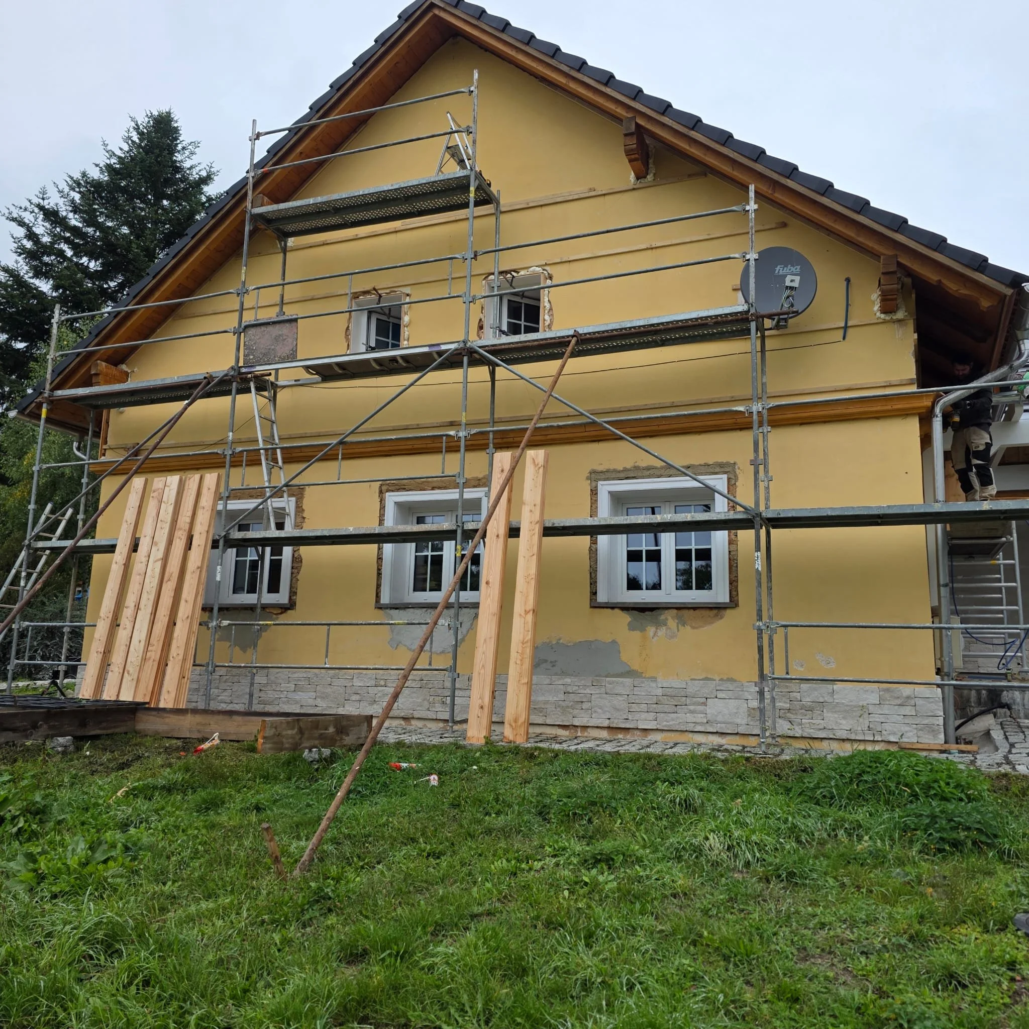 A house under renovation with scaffolding surrounding it. The house has a yellow exterior with three windows on the lower level and two on the upper level. There are wooden planks leaning against the house and a person visible on the right side of th