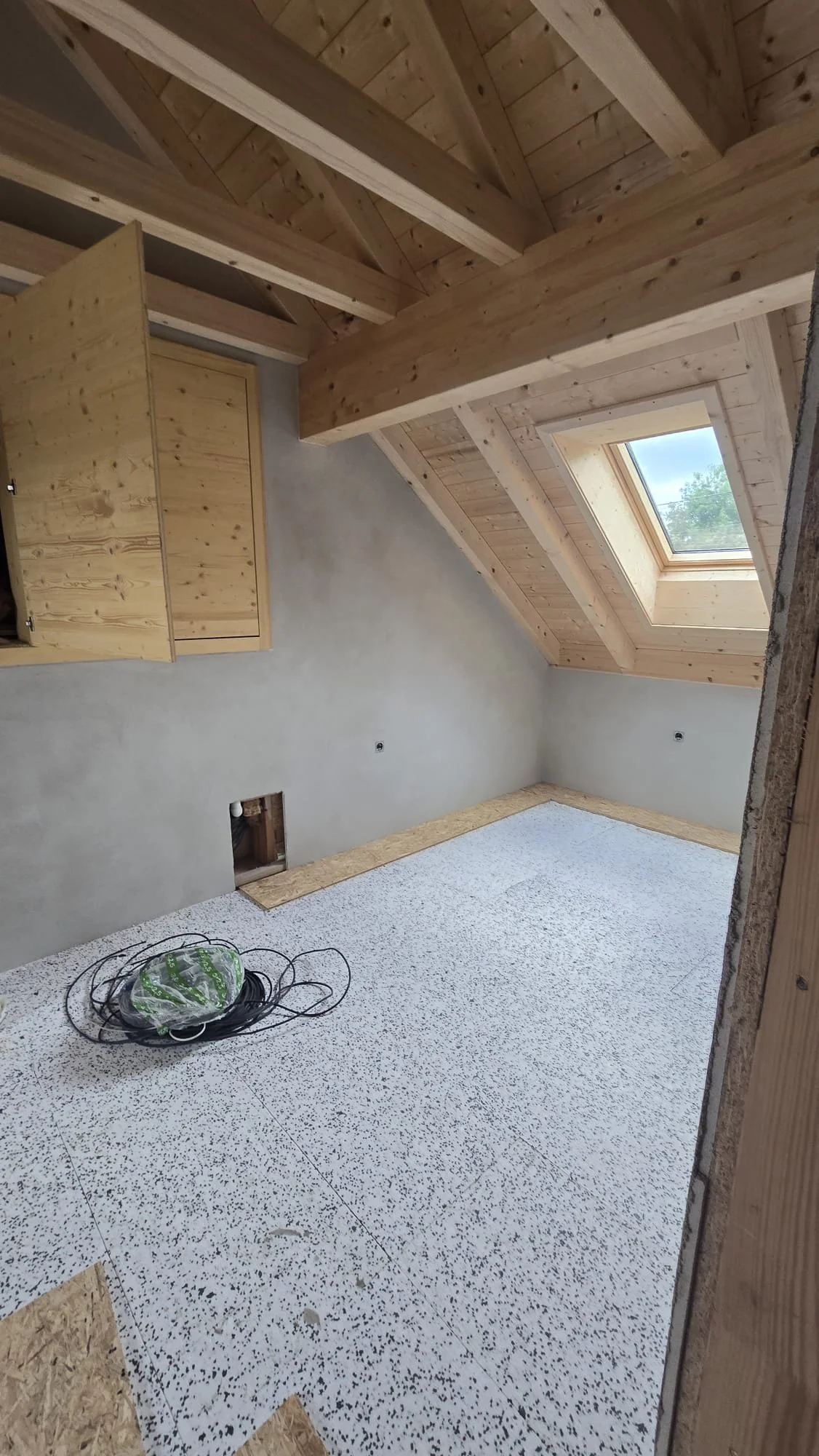 An unfinished attic room with wooden beams and a skylight window, with a partially installed floor and a bundle of electrical wires on the floor.