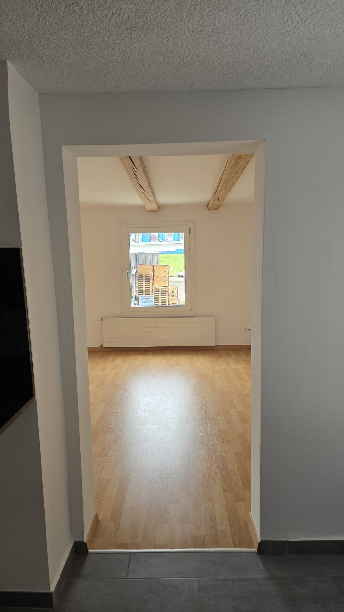 Empty room with a window, wooden beams on the ceiling, and light-colored flooring.