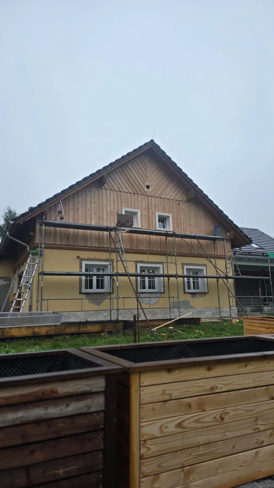 A house under construction or renovation with scaffolding set up around it. The house has a wooden upper section and a yellow lower section with three windows. There is construction equipment and materials in the foreground.