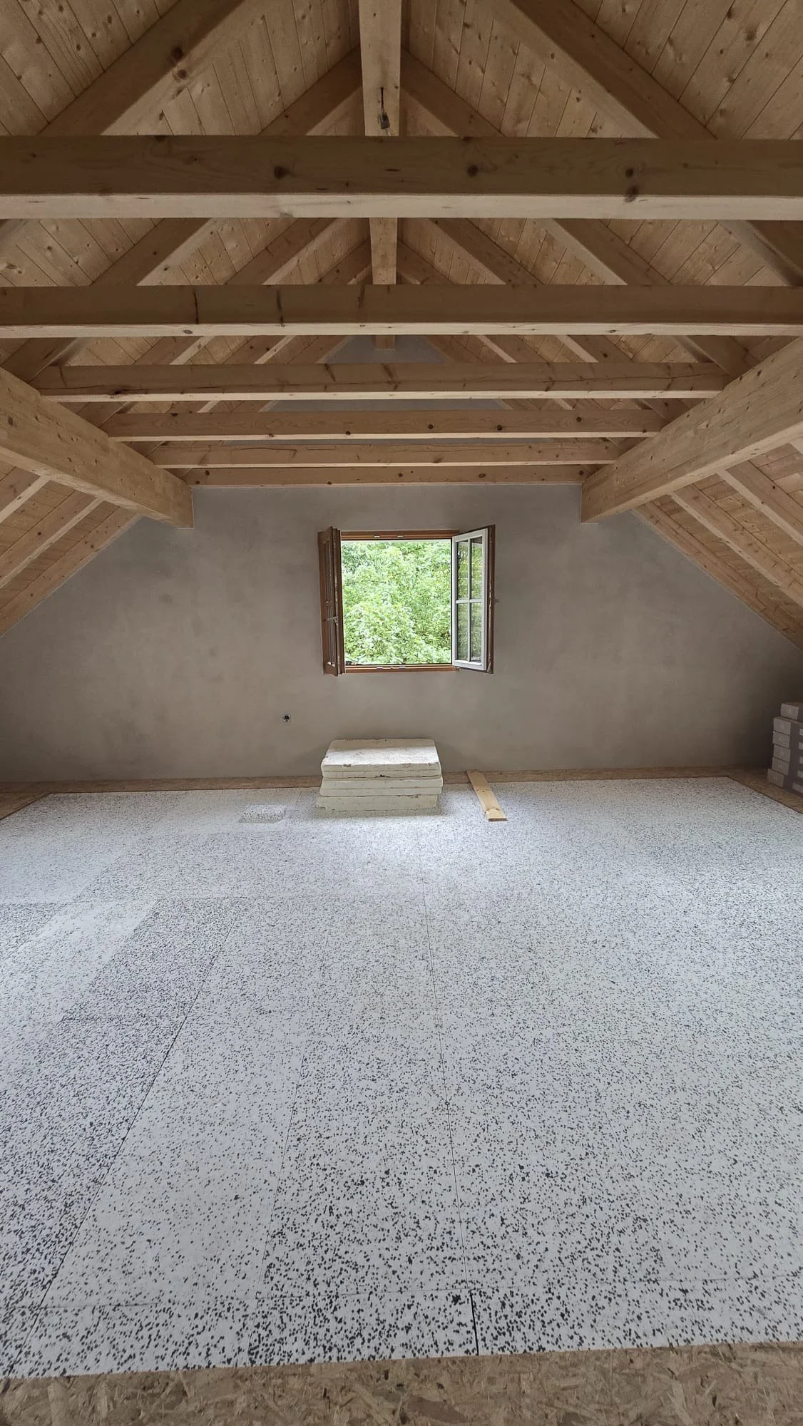 An unfinished attic room with a wooden ceiling and exposed beams, a small open window showing green trees outside, and a speckled flooring. There are some construction materials on the floor.