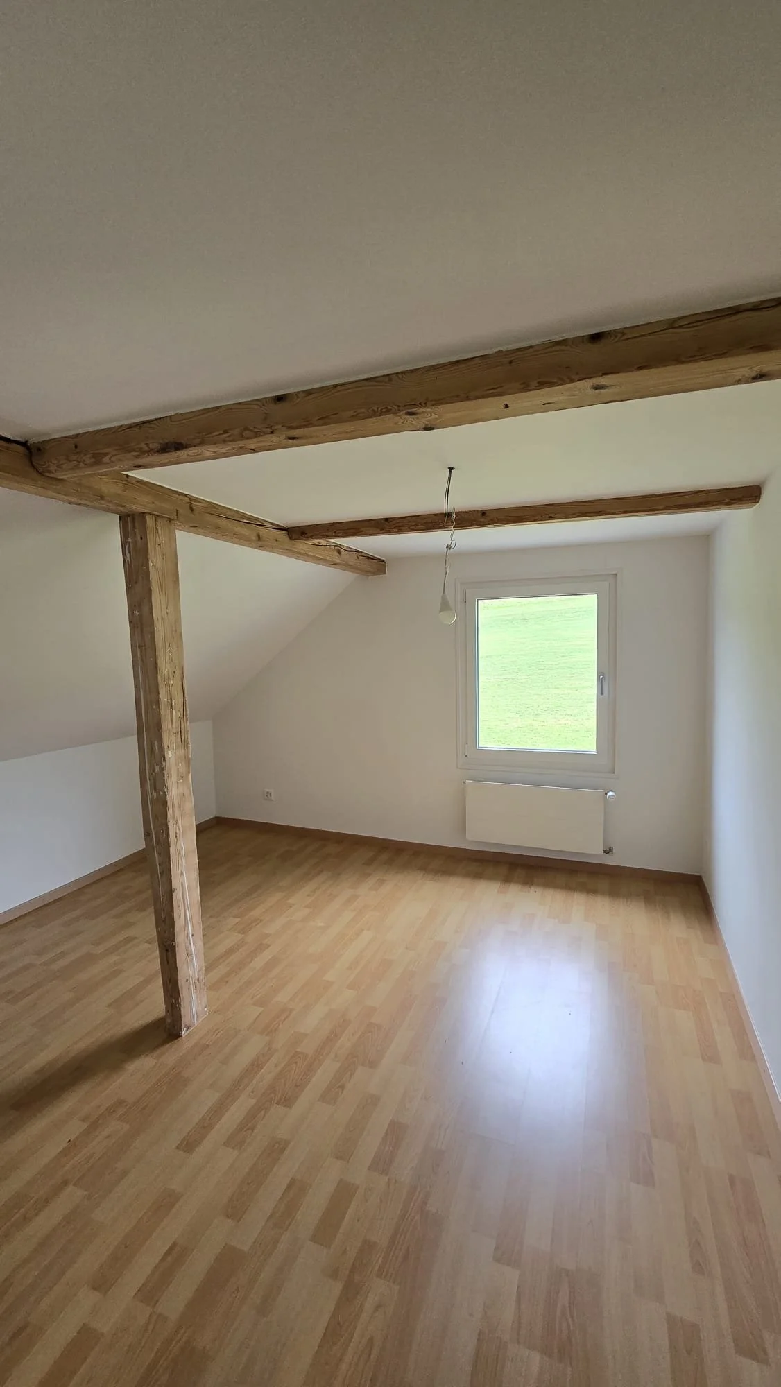 Empty attic room with wooden beams, a window showing green grass outside, and light wood flooring.