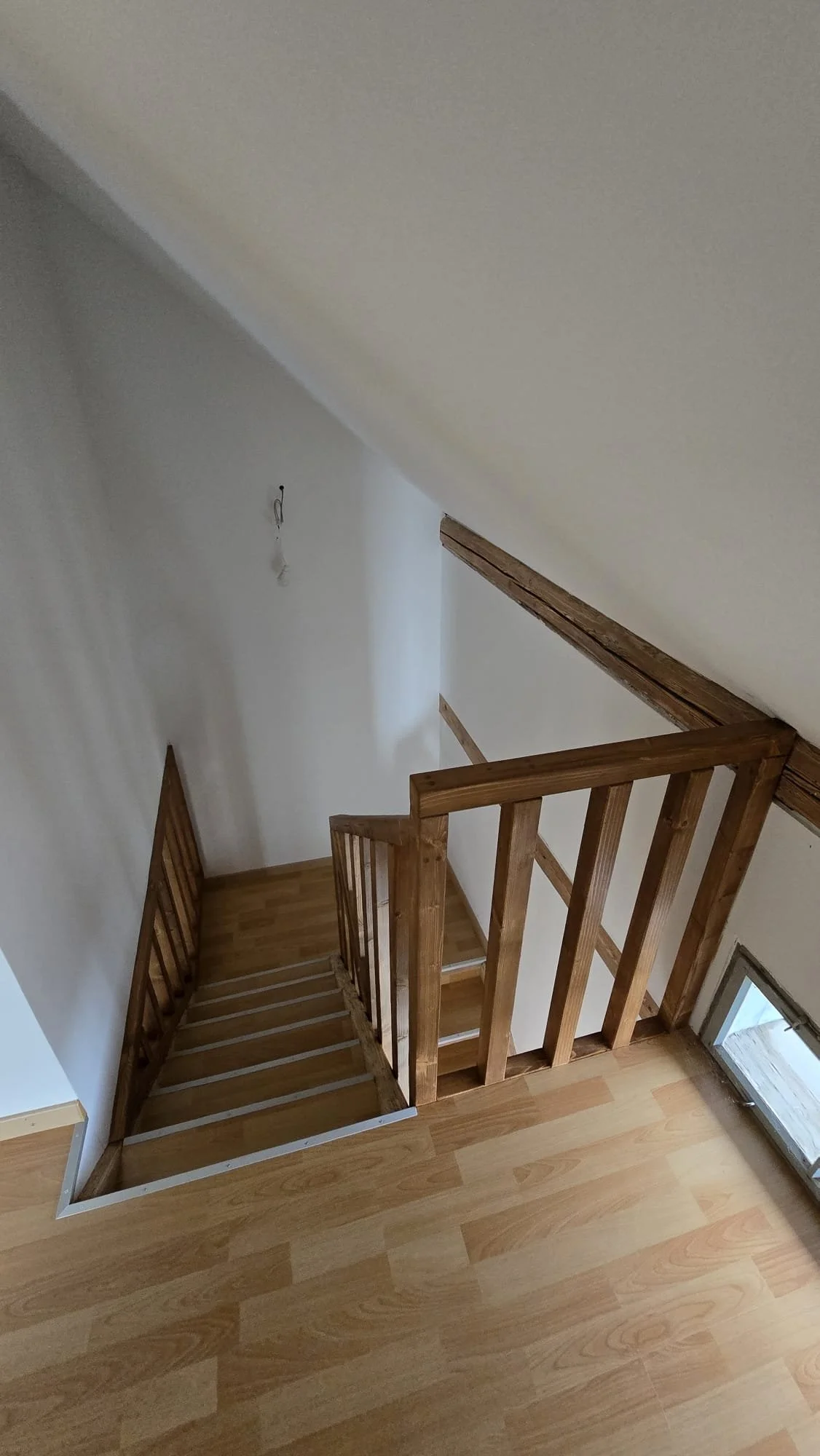 View from the top of a wooden staircase leading down to an uncarpeted landing with white walls and a small window at the bottom.