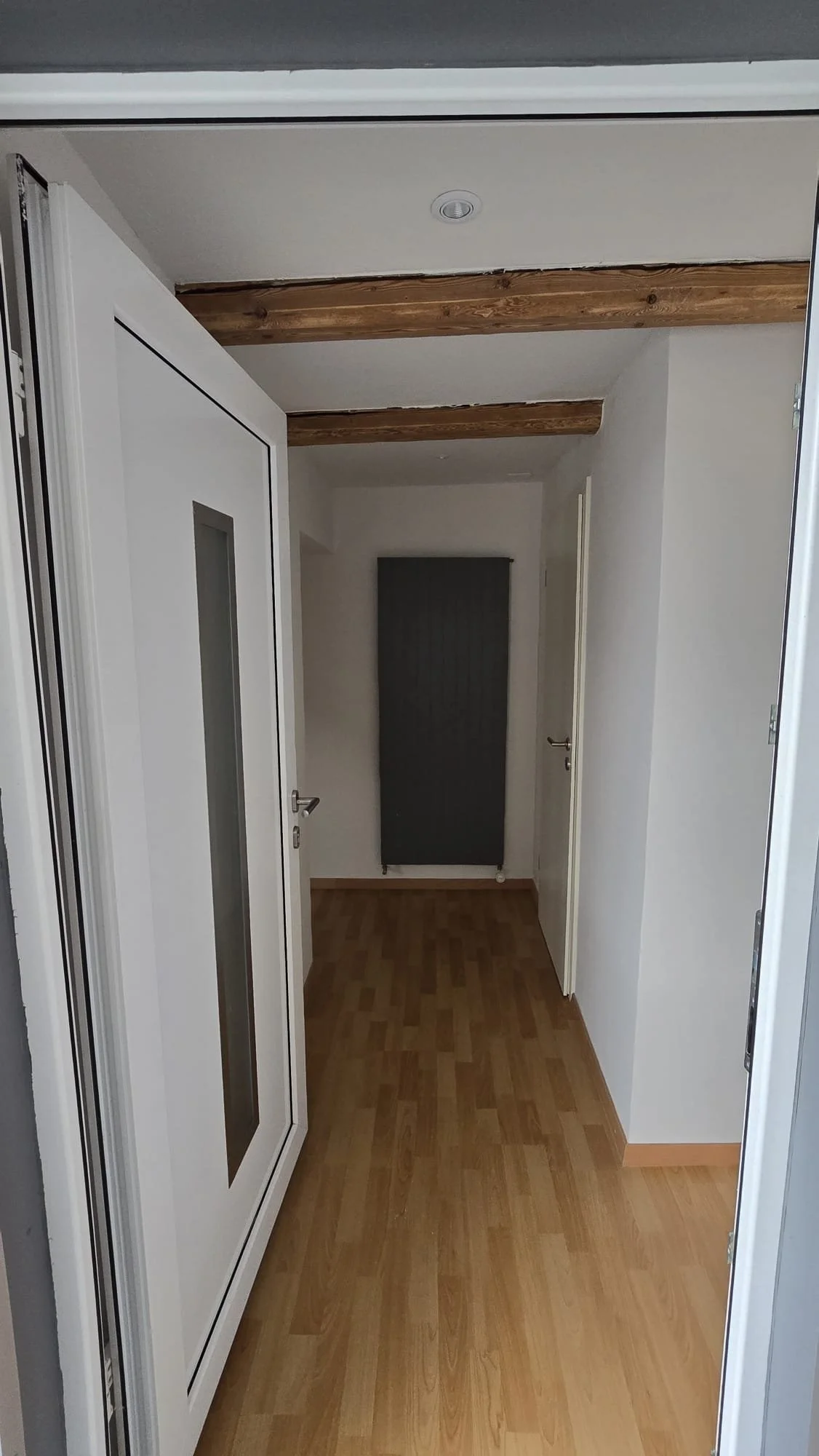 Empty hallway with wooden beams on the ceiling, white walls, a black radiator on the far wall, and a white door on the right side.