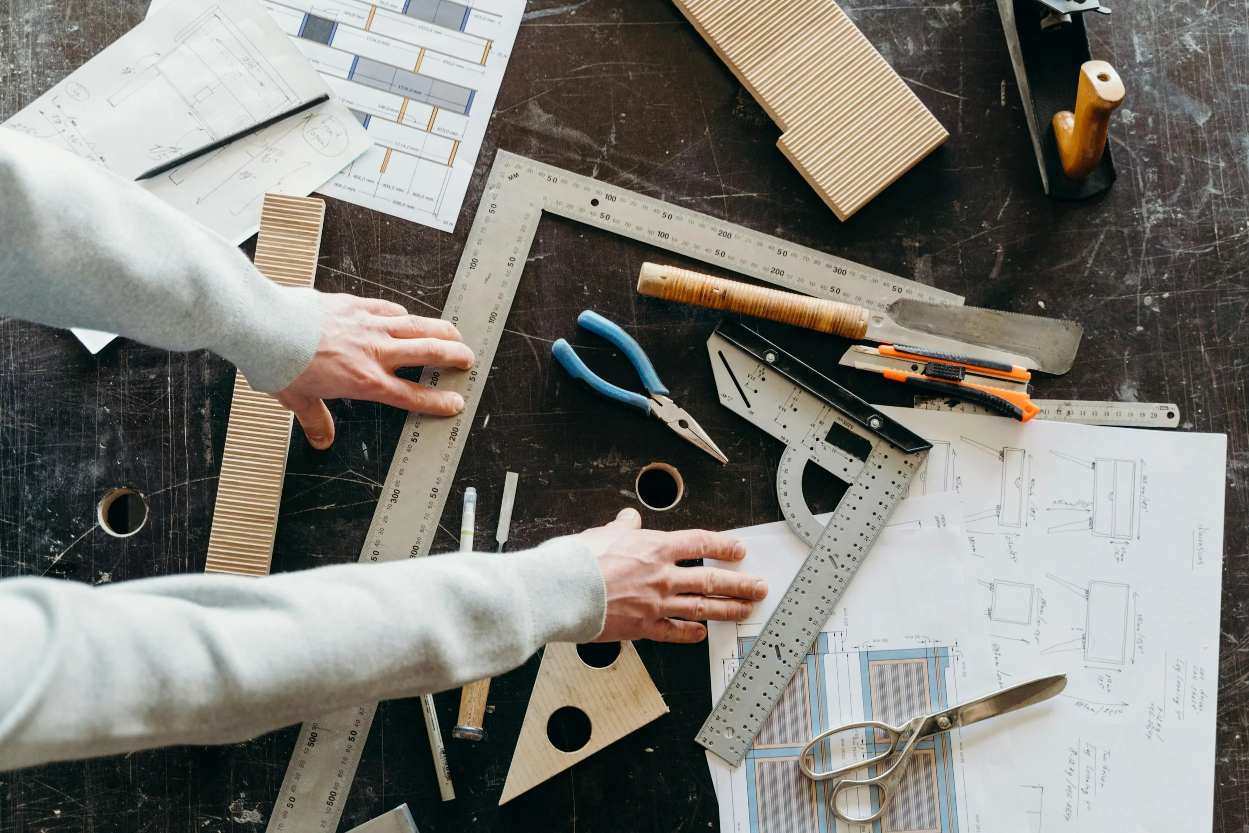 Overhead view of a work desk with drafting tools, blueprints, and sketches, where a person is measuring with a ruler.