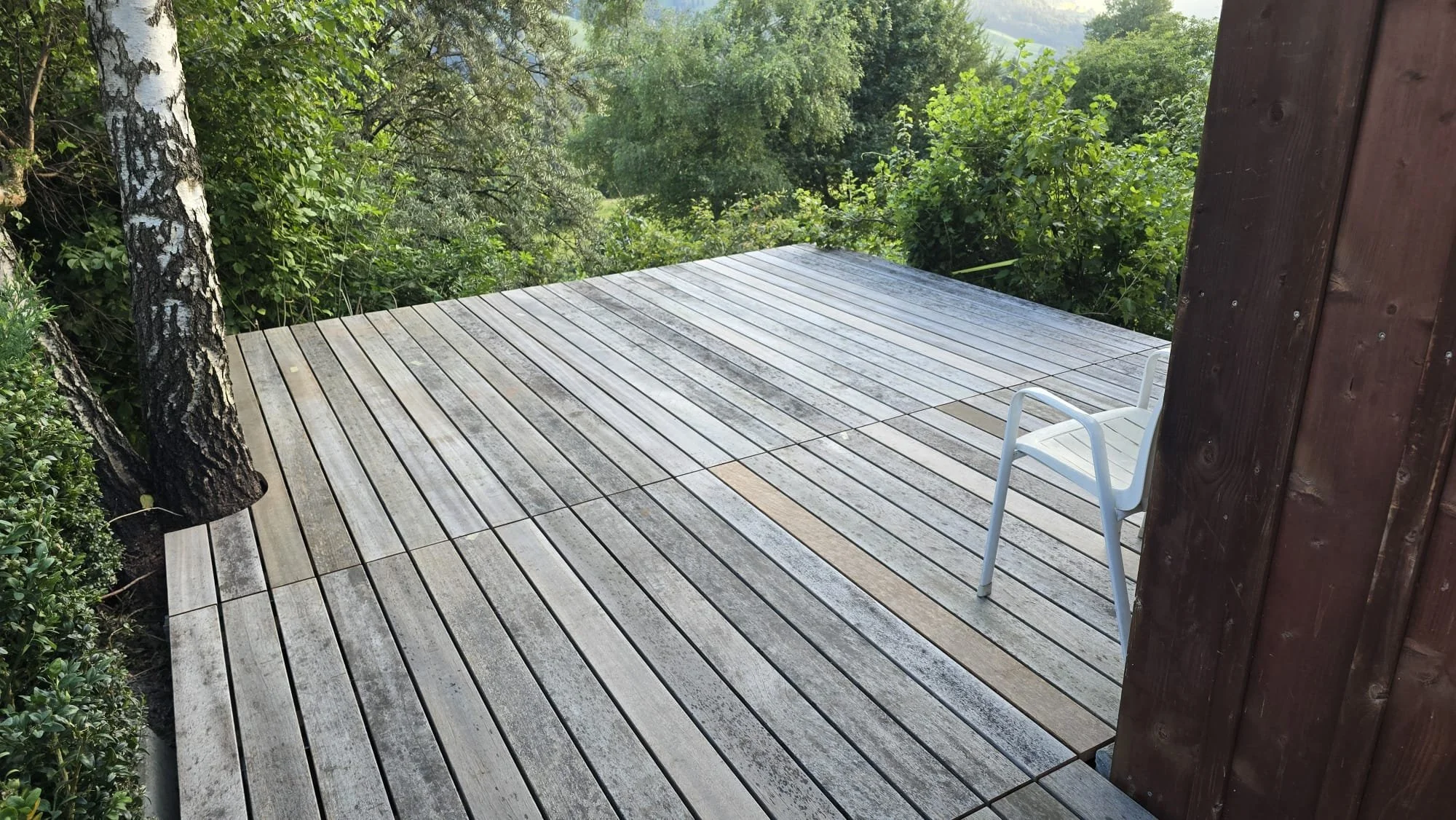 A wooden deck with weathered planks surrounded by green trees and bushes, with a white plastic chair against a dark wood post on the right side.