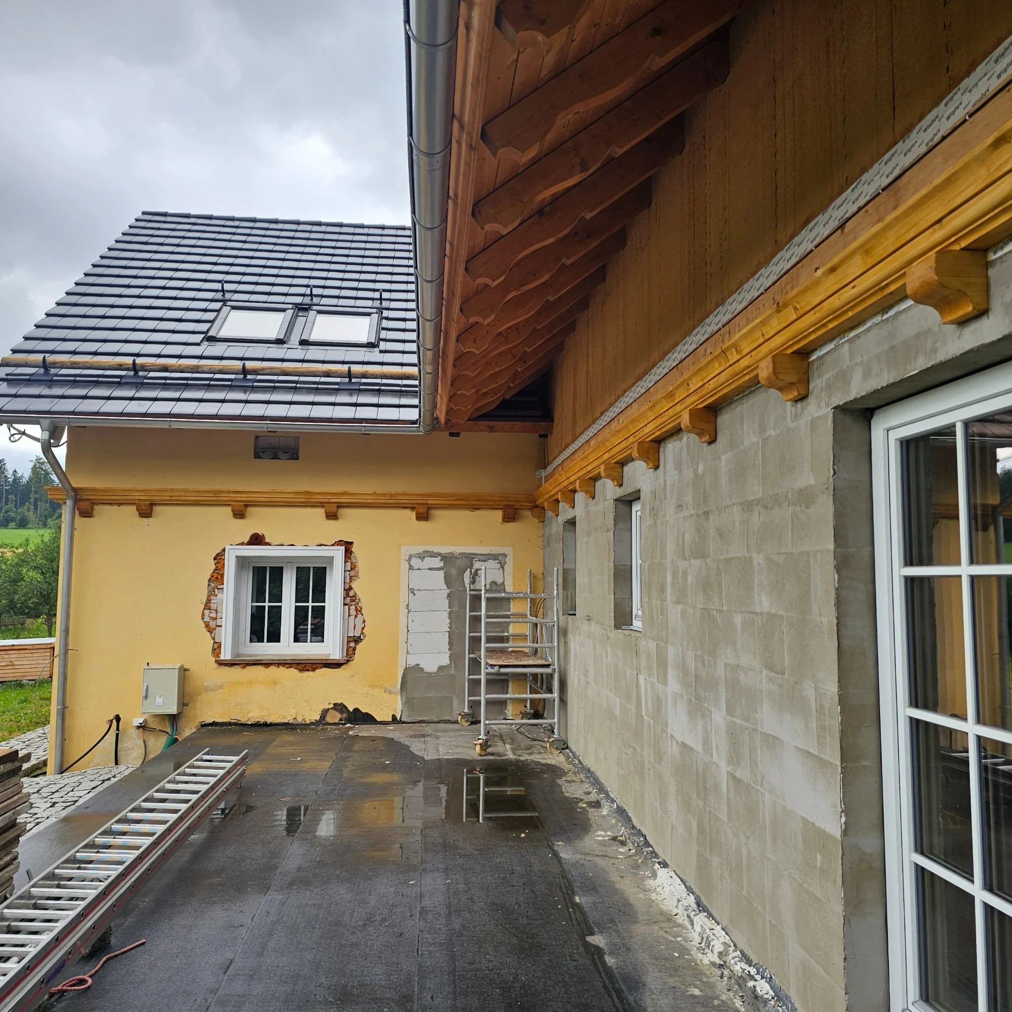Construction site of a house exterior with scaffolding, a partially finished wall, window with decorative brick border, and a tiled roof with skylight in cloudy weather.