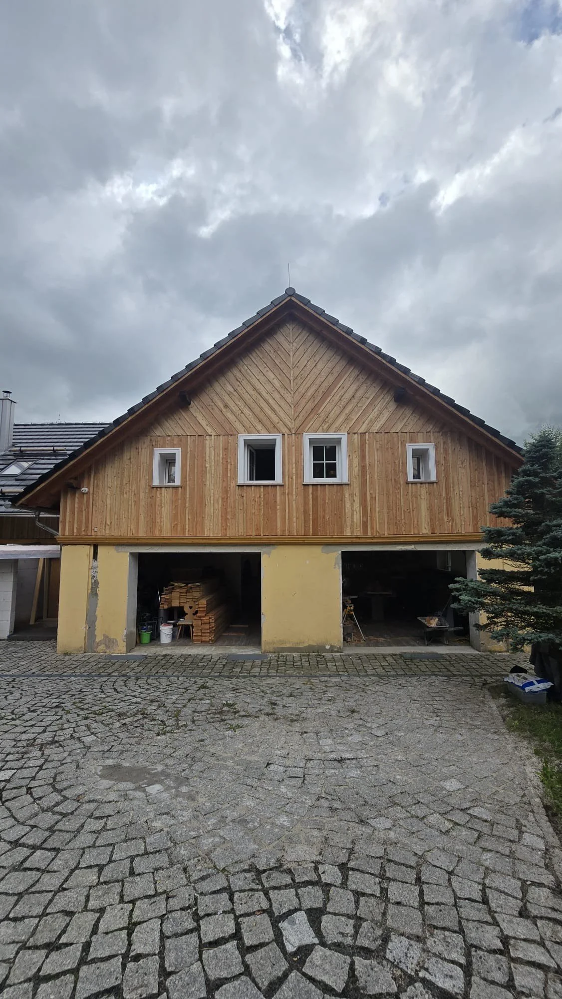 A house under construction with a wooden upper floor and a yellow lower floor, with open garage spaces showing construction materials inside, on a cobblestone driveway under a cloudy sky.