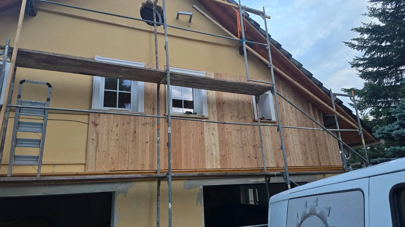 House under construction with scaffolding and new wooden siding being installed, and a partial view of a white utility vehicle in the foreground.