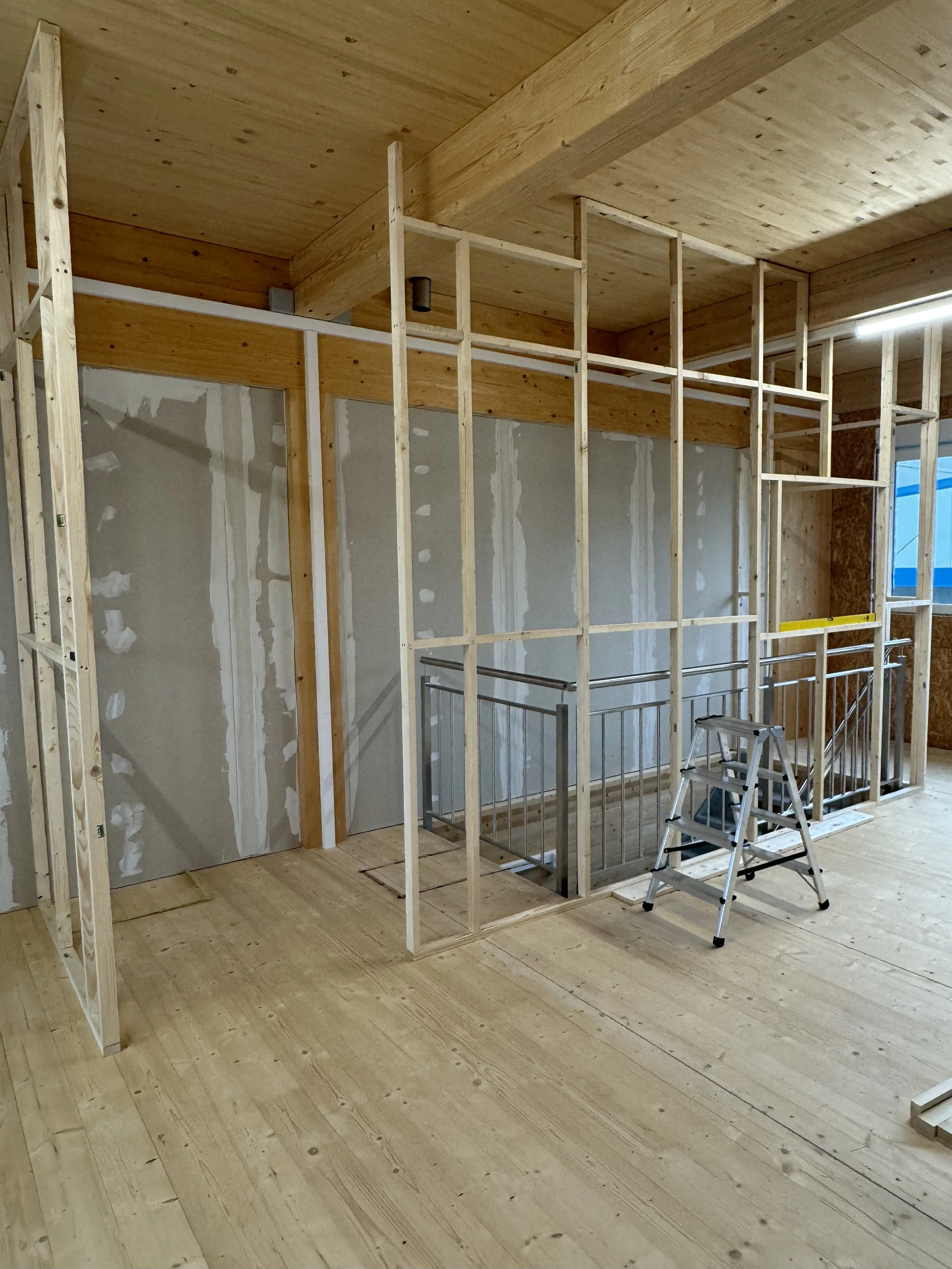 Interior of a building under construction with wooden framing and staircase, unfinished drywall on the wall, and a small step ladder.