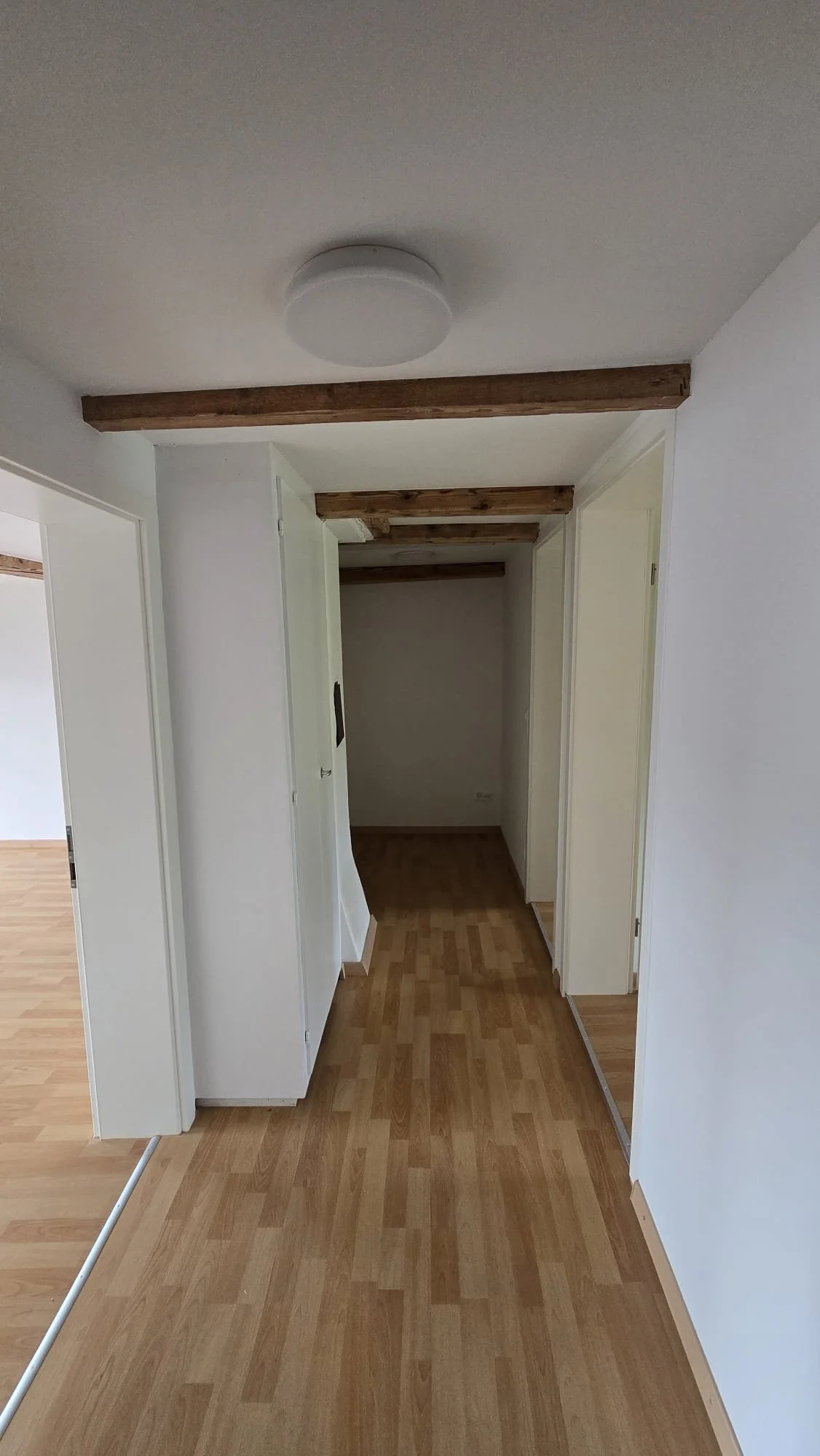 Empty hallway with wood flooring and white walls, wooden beams on the ceiling, and a doorless doorway leading to an adjacent room.