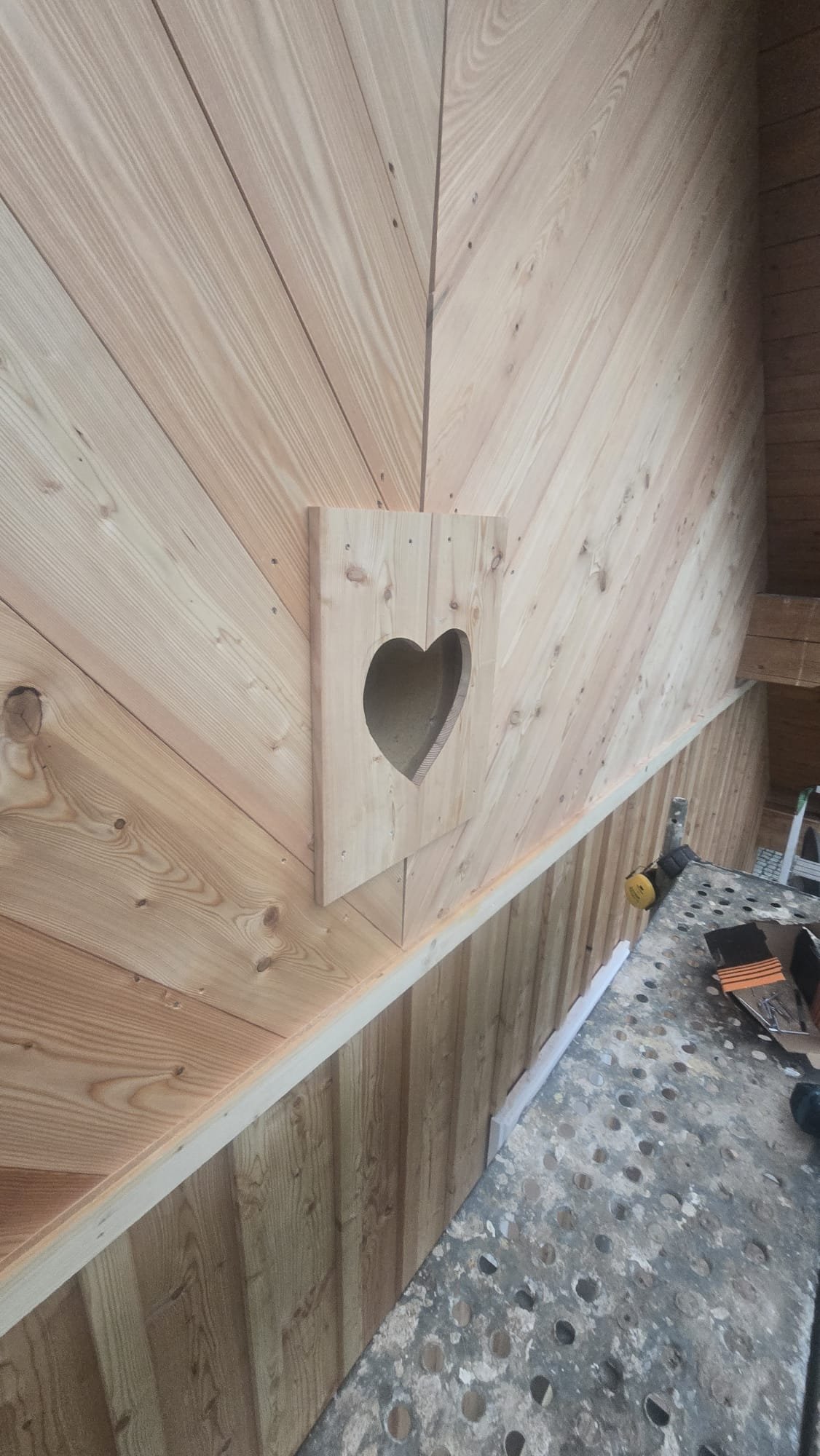 Wooden shelf with a heart-shaped cutout on a wood-paneled wall, in a workshop with tools and a workbench.