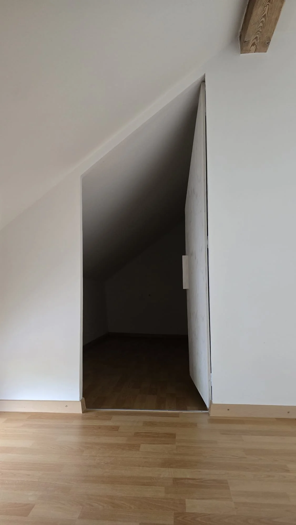 Interior of a room with light-colored wooden flooring, a doorway with a door slightly ajar leading into a dark, small attic or storage space under a sloped ceiling.