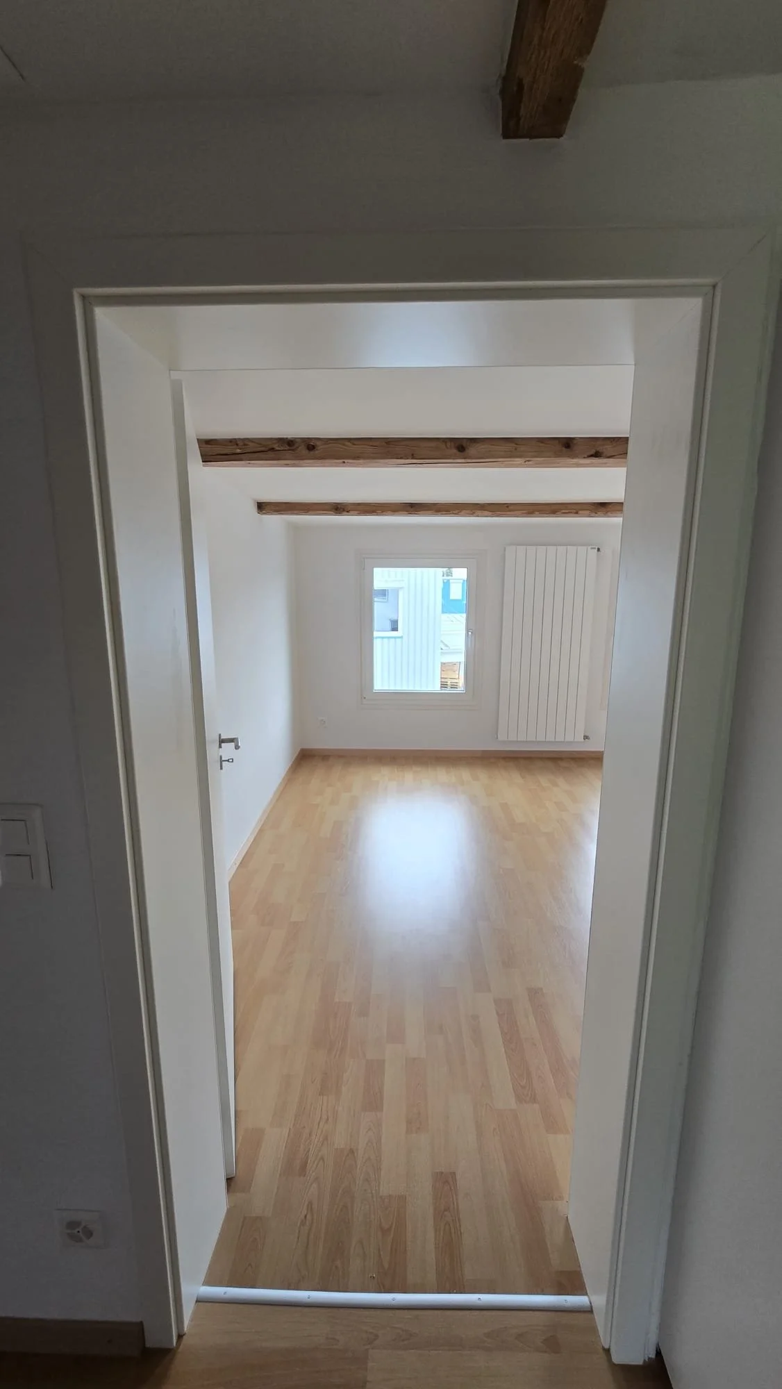 Empty room with wooden beams on the ceiling, a window, and a radiator, viewed from a doorway.